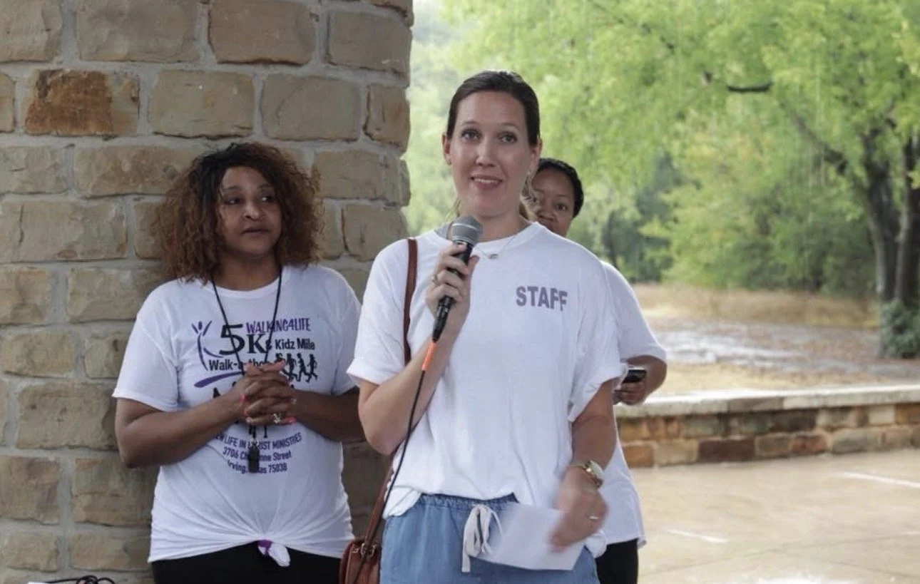 A woman holding a microphone and speaking at an outdoor event, with two women standing behind her near a brick structure, surrounded by green trees.