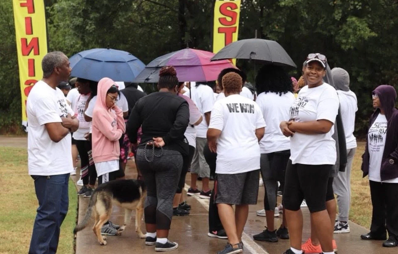 A group of people gathered outdoors on a rainy day, some holding umbrellas. They are dressed in white T-shirts, likely participating in a commemorative event. One person is smiling at the camera, and there is a German Shepherd dog in the scene. Yellow banners with red text are visible in the background.