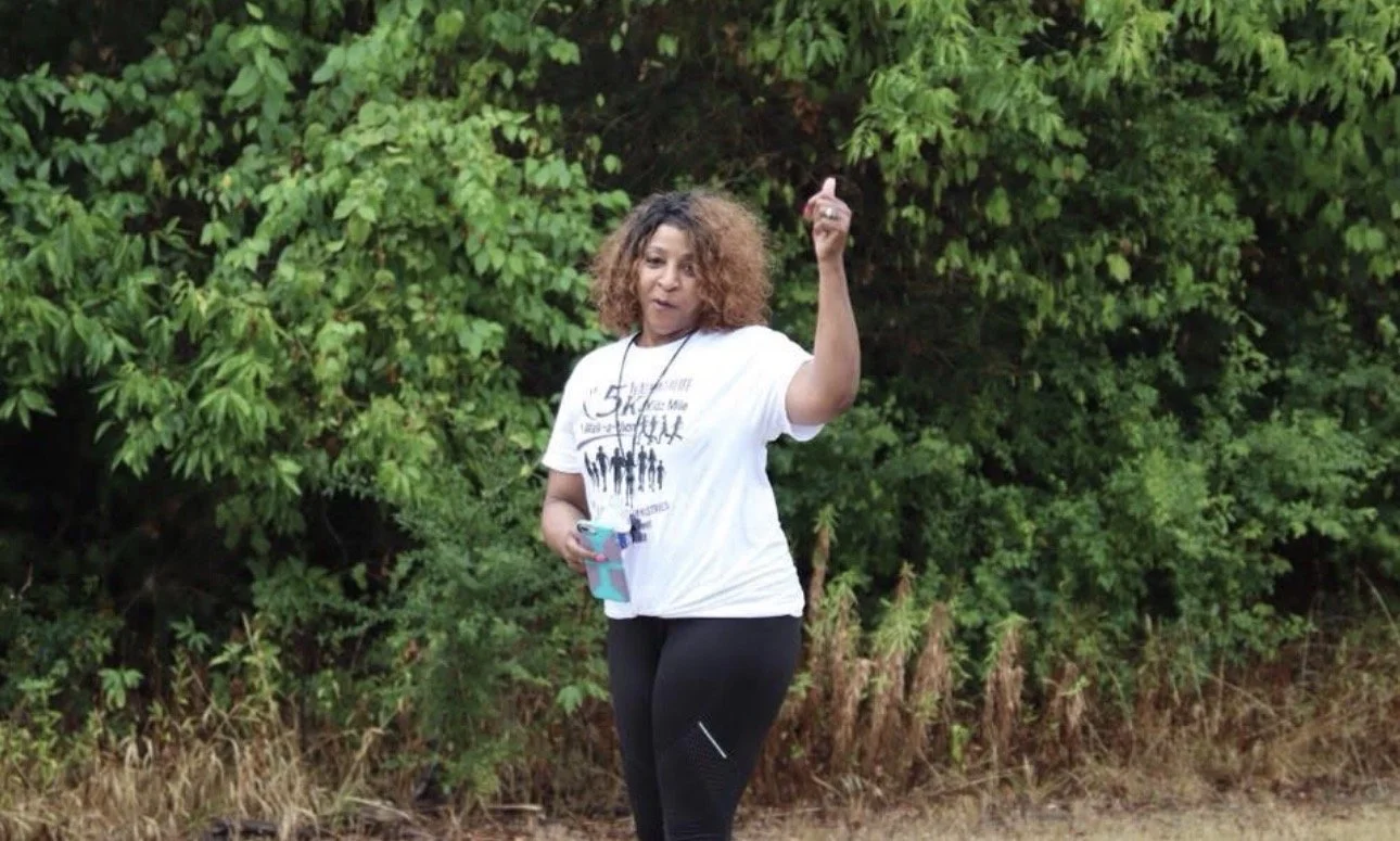 Woman with curly hair wearing a white t-shirt and black leggings standing outdoors near green bushes, holding a phone in her left hand and pointing upwards with her right hand.