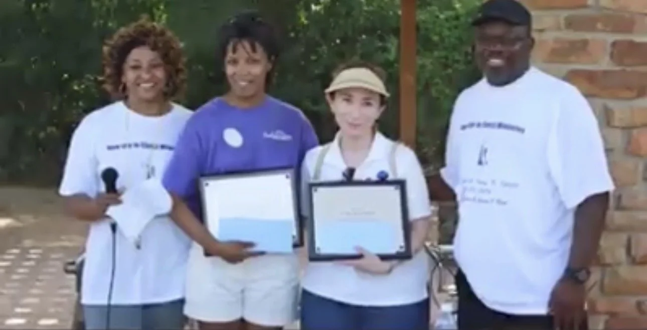 Four people outdoors, two children holding certificates, one woman holding a microphone, one man standing beside them, all smiling.