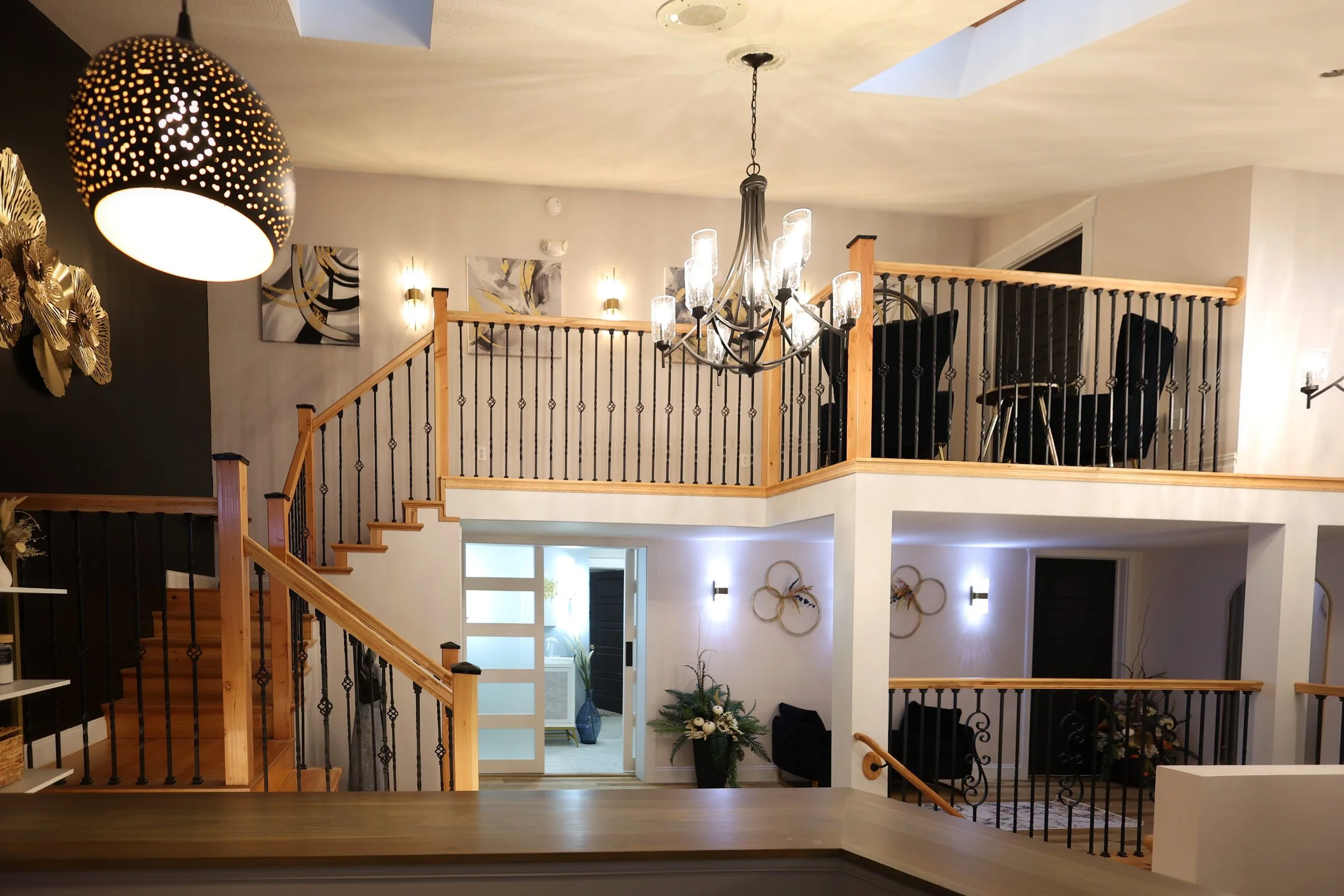 Interior view of a multi-level home with a staircase, featuring decorative wall art, a large hanging chandelier, and a wall lamp, with a small plant and additional seating area visible on the lower level.