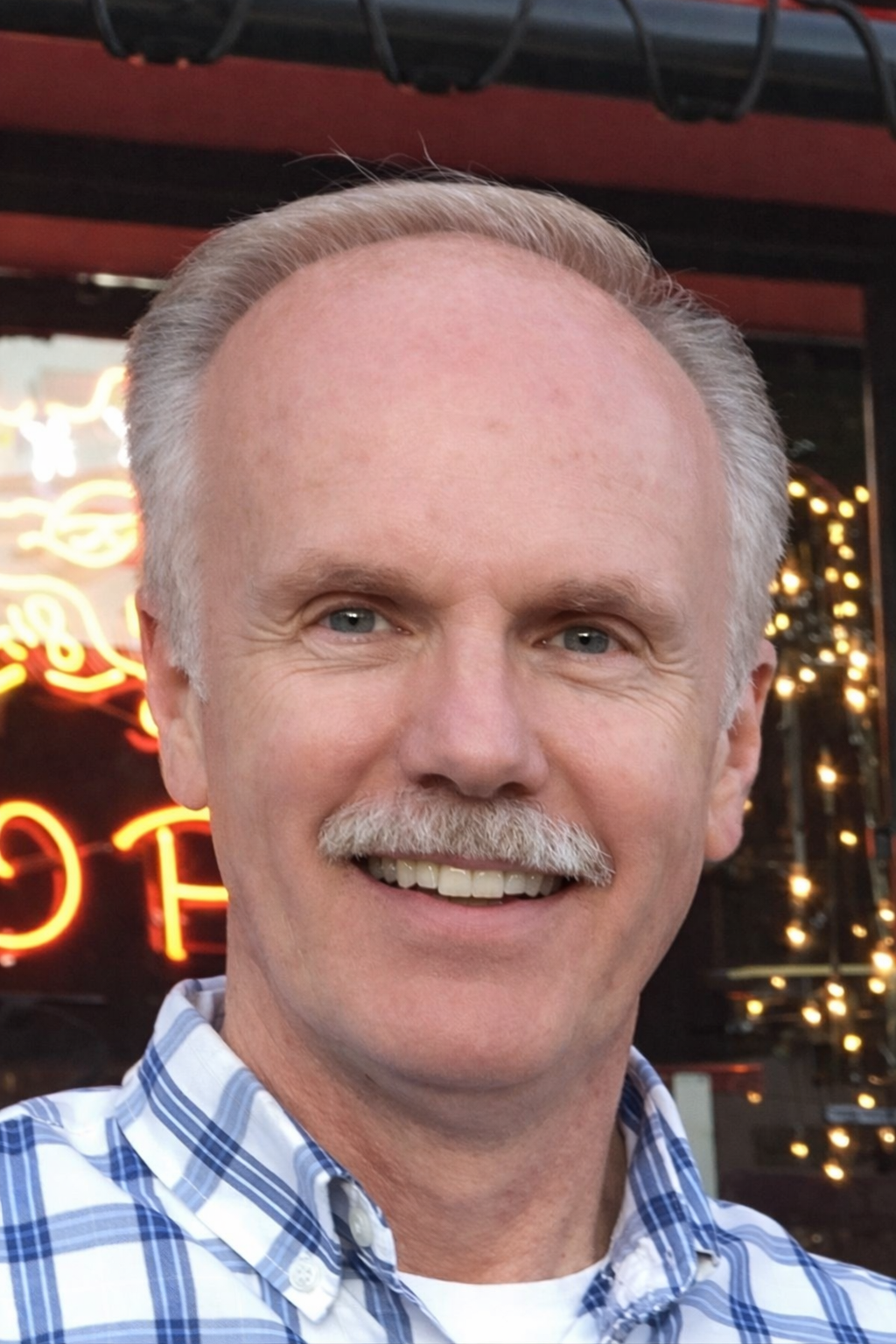 A smiling middle-aged man with white hair and a mustache, wearing a blue and white checkered shirt, standing outside with neon lights visible in the background.