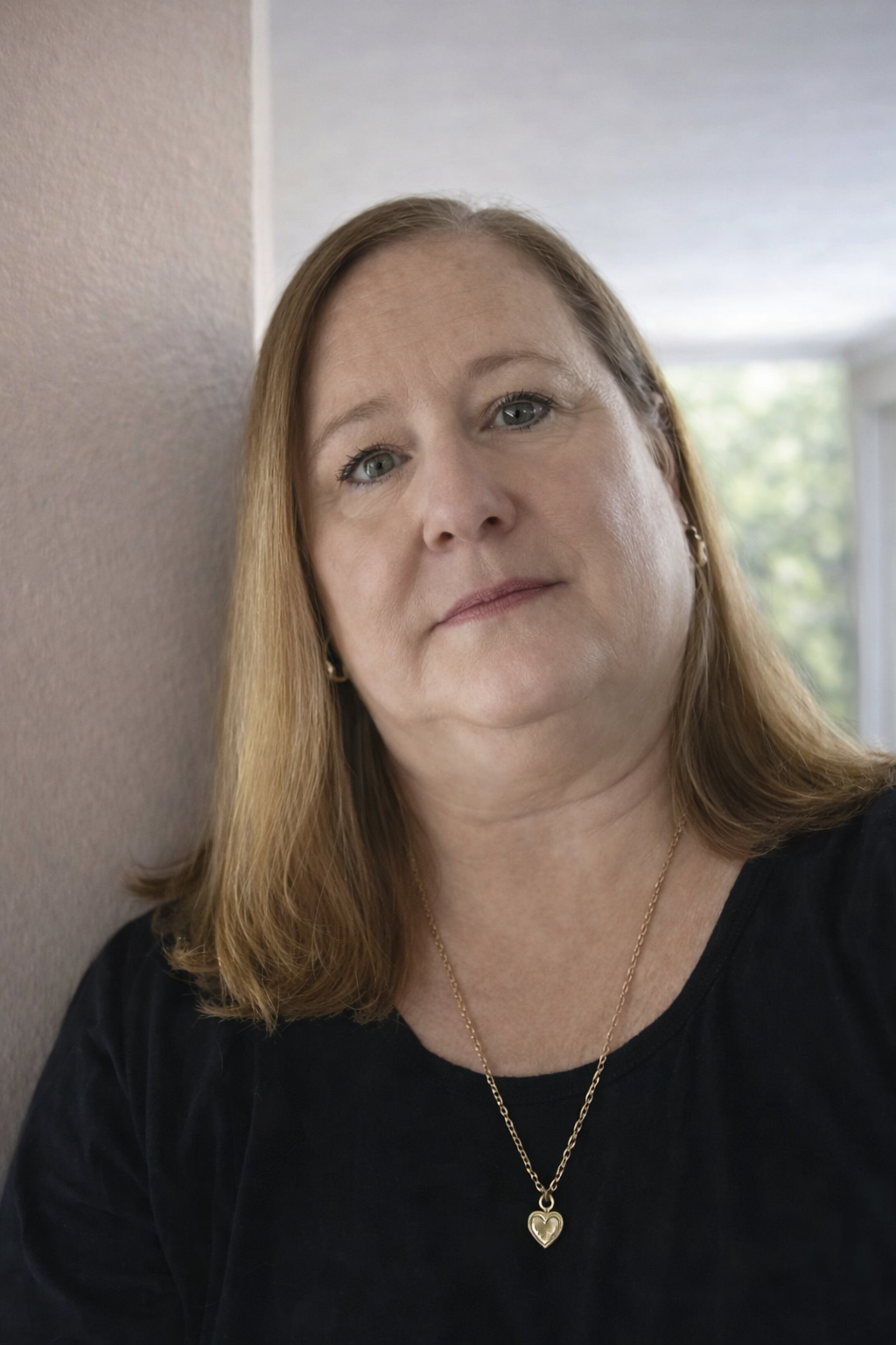 A woman with shoulder-length light brown hair posing indoors against a wall, wearing a black top and a gold necklace with a heart-shaped pendant.