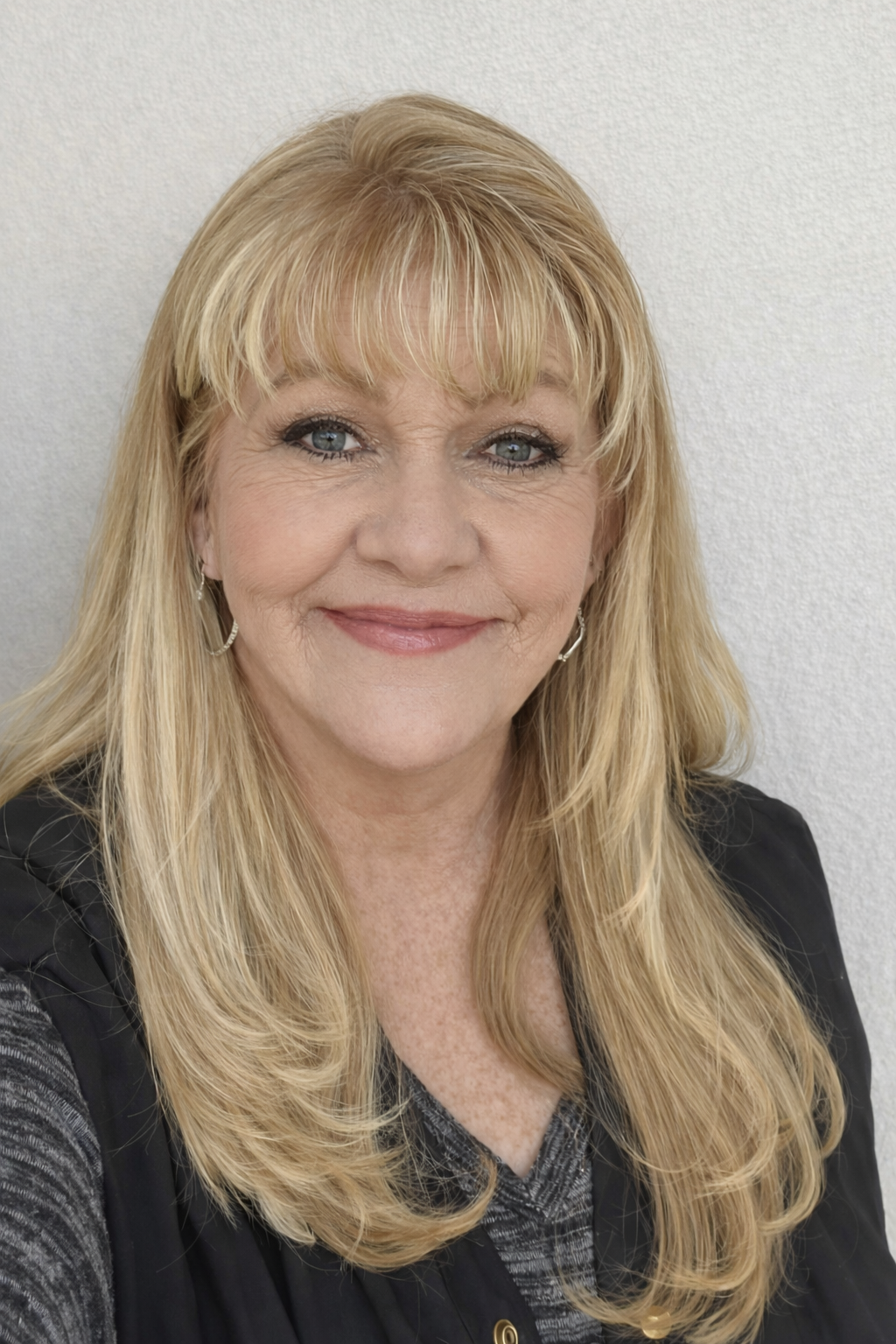 A woman with long blonde hair, blue eyes, and earrings, smiling at the camera against a plain white background.