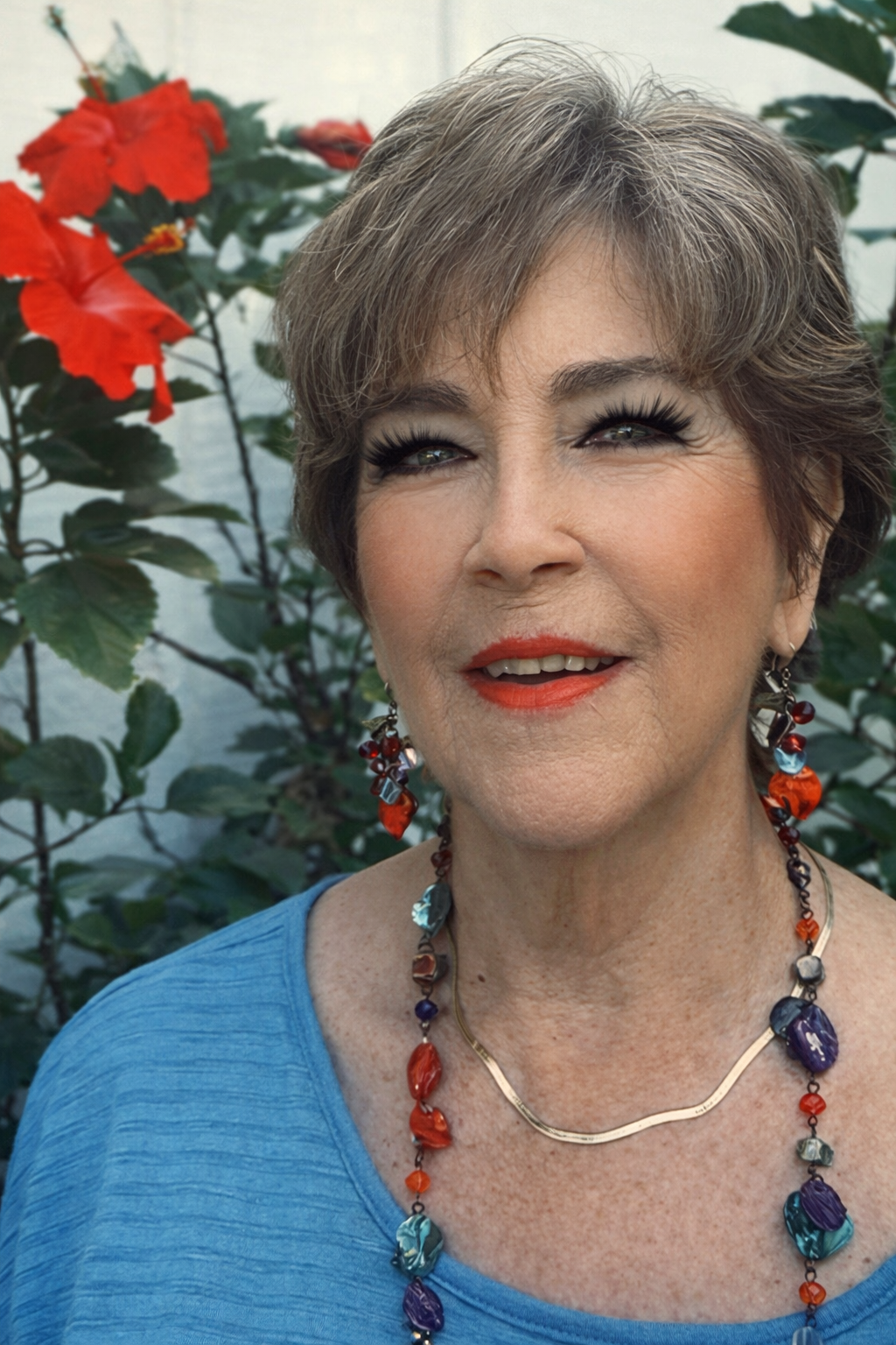 A woman with short gray hair, wearing a blue top, colorful beaded jewelry, and red lipstick. She is outdoors with red hibiscus flowers and green leaves in the background.