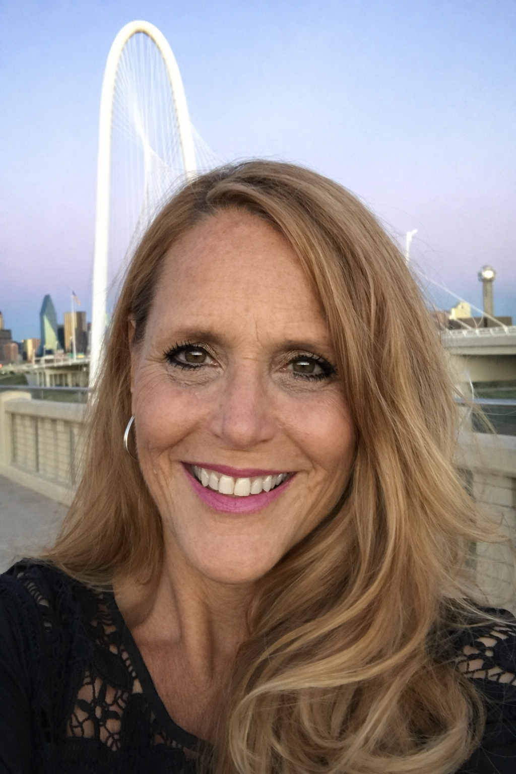 Close-up of a smiling woman with long, wavy blonde hair, wearing makeup and a black lace top, in front of the Dallas skyline with the giant white observation wheel, the Margaret Hunt Hill Bridge, and the cityscape at sunset.