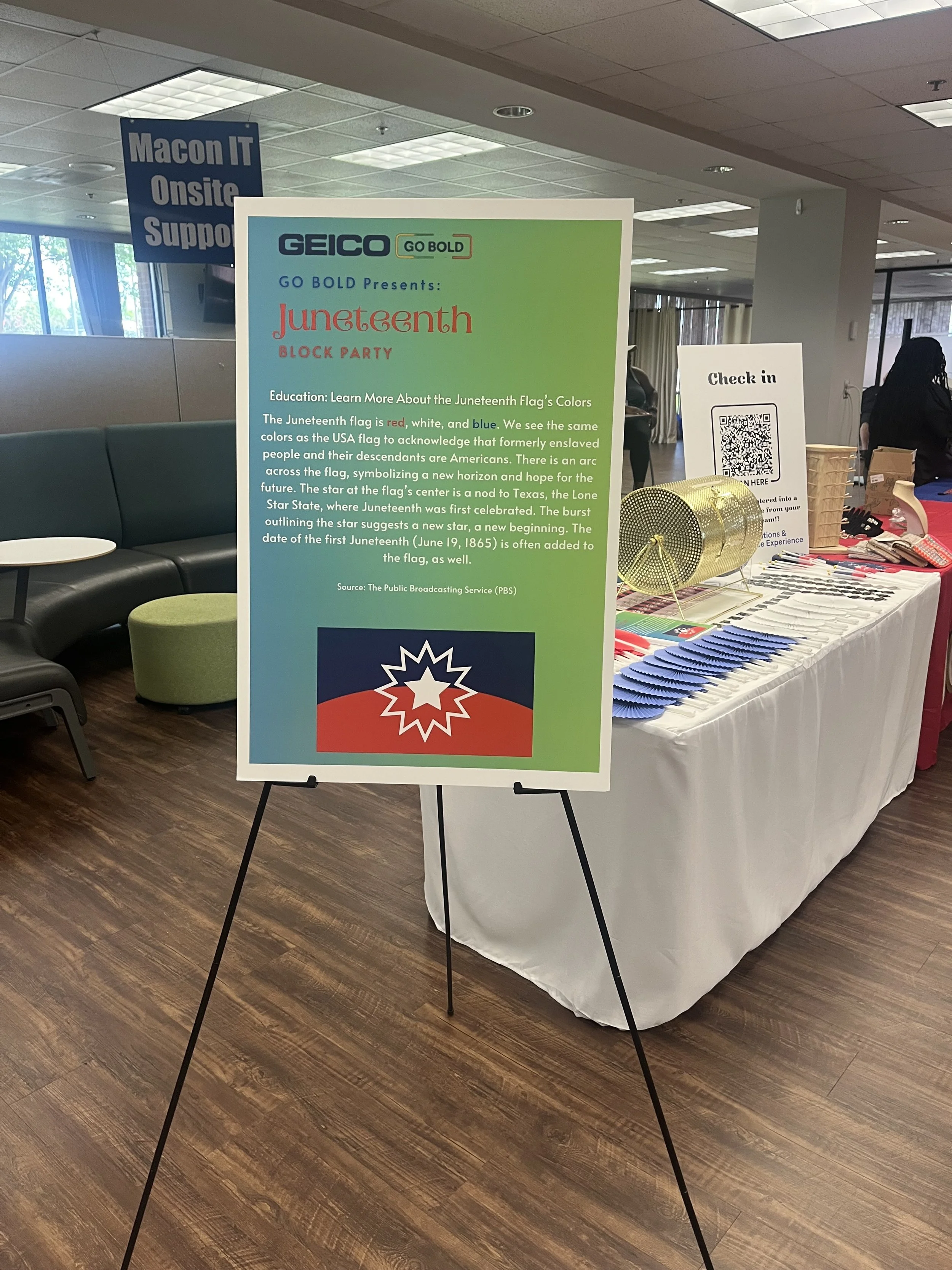Sign promoting a Juneteenth block party event by GEICO, with information about Juneteenth flag colors and their meaning, and a QR code for check-in, placed on a stand in an indoor event space with tables displaying items and a banner in the background.