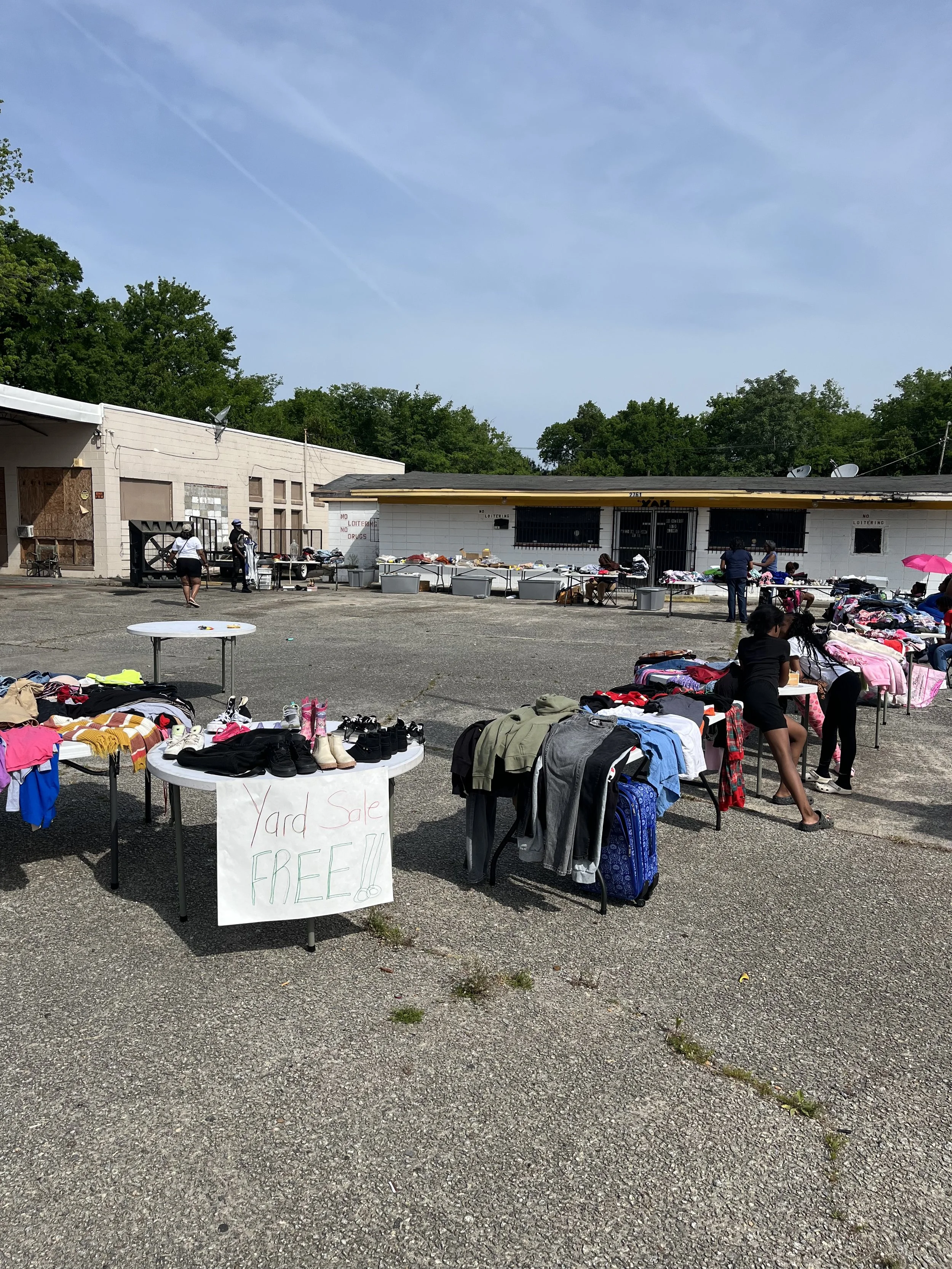 An outdoor yard sale with tables displaying clothing, shoes, and other items. A handwritten sign reads 'Yard Sale FREE!!'. Several people are browsing and shopping, with some sitting at the tables. Buildings and trees are in the background under a partly cloudy sky.