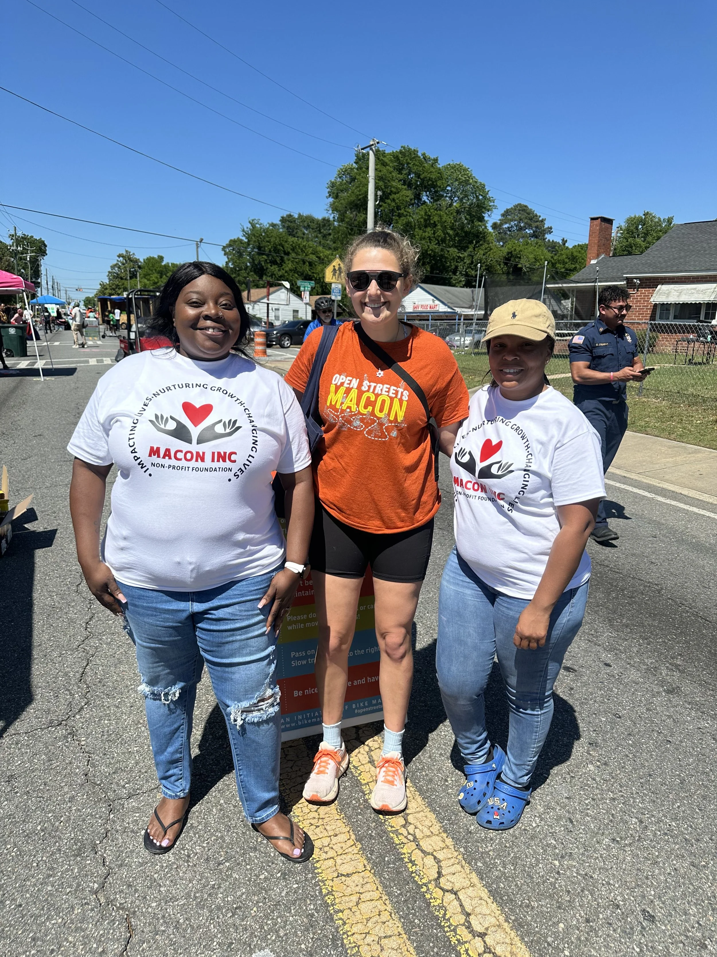 Three women standing on a street, smiling at the camera during a community event on a sunny day. Two women wear white T-shirts with the logo 'Macon Inc,' and the third woman wears an orange T-shirt with the words 'Open Streets Macon.' There is a police officer in the background and tents set up along the street.