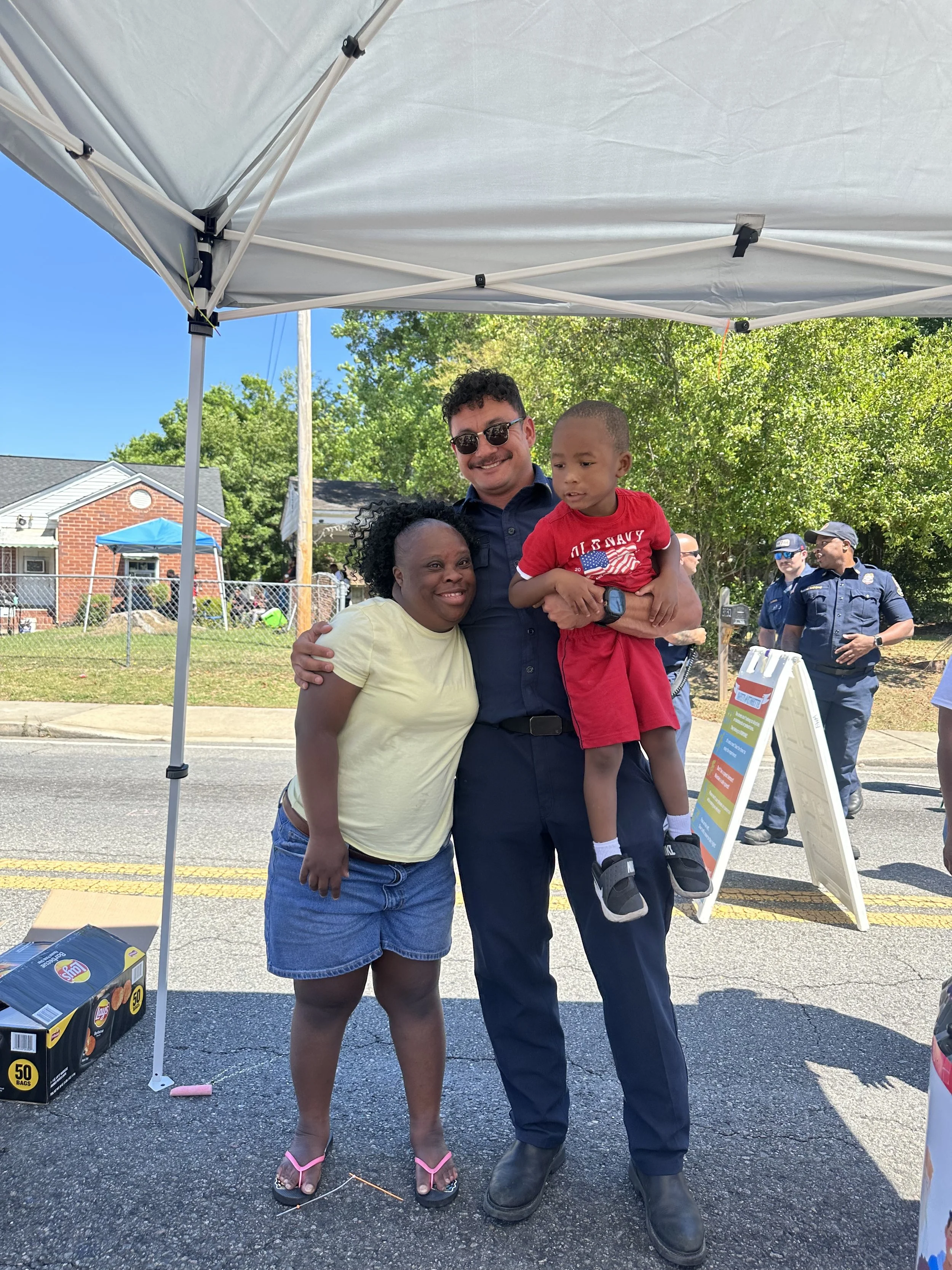 Three people are standing together outdoors under a canopy. A woman with dark, curly hair is on the left, smiling, wearing a pale yellow shirt and shorts with pink flip-flops. A man with sunglasses is in the middle, smiling, wearing a dark blue uniform, and holding a young boy dressed in a red shirt and black shorts. In the background, there are police officers and a small crowd near a chain-link fence, with trees and houses visible.