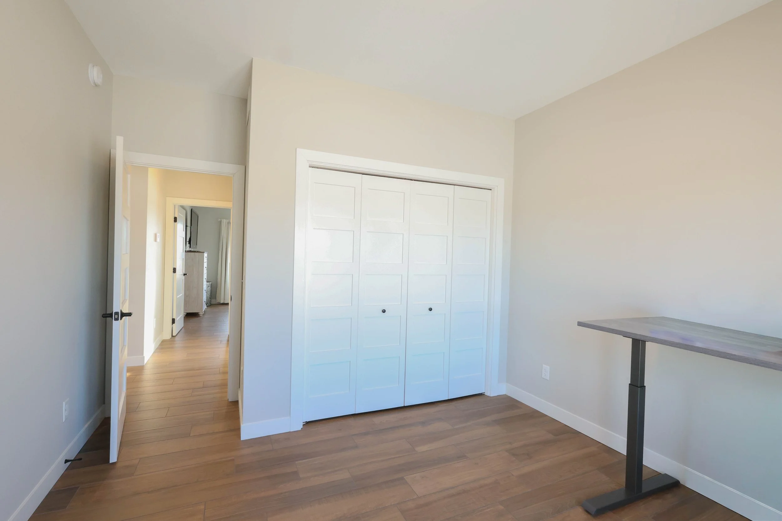 Empty room with light-colored walls, hardwood floors, a white sliding closet door, a partially open door leading to another room, and a small table or desk near the right edge.