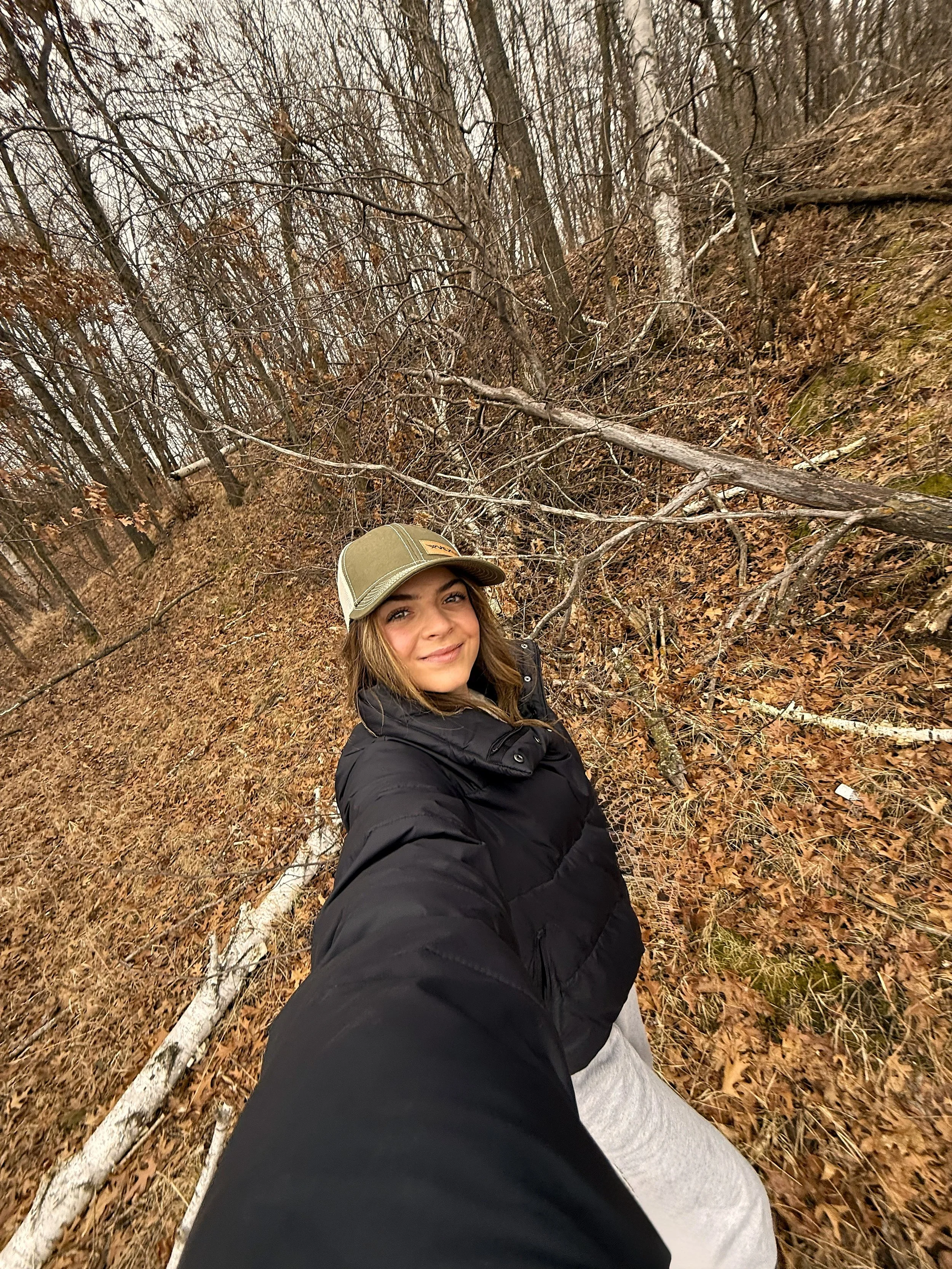 A young woman with brown hair, wearing a green cap and black jacket, smiling while taking a selfie in a wooded area with fallen leaves and leafless trees.