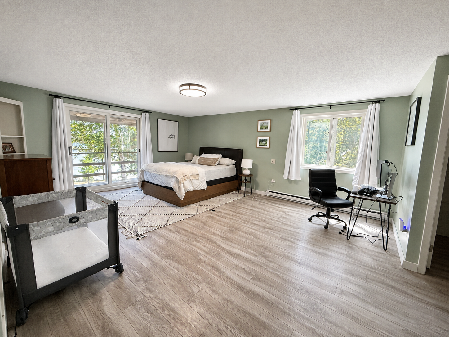 A spacious bedroom with green walls, wood flooring, a bed with dark headboard, nightstand with lamp, window with white curtains, small desk with computer and office chair, and sliding glass door leading outside.