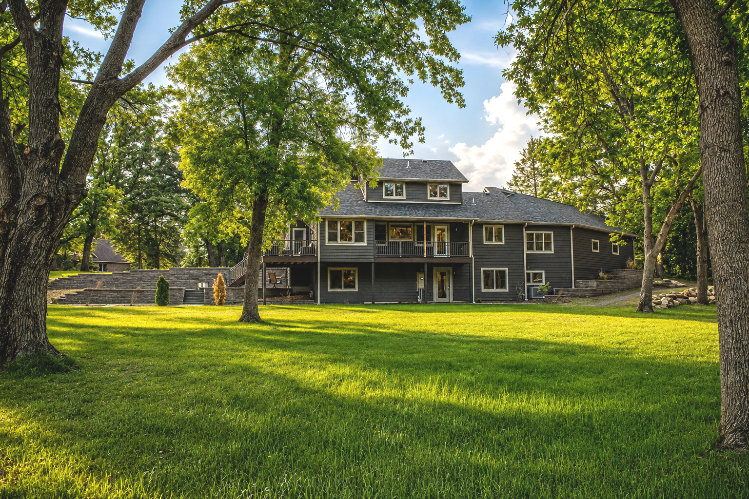 Large house with multiple windows and a backyard deck, surrounded by lush green trees and grass, under a partly cloudy sky in a suburban setting.