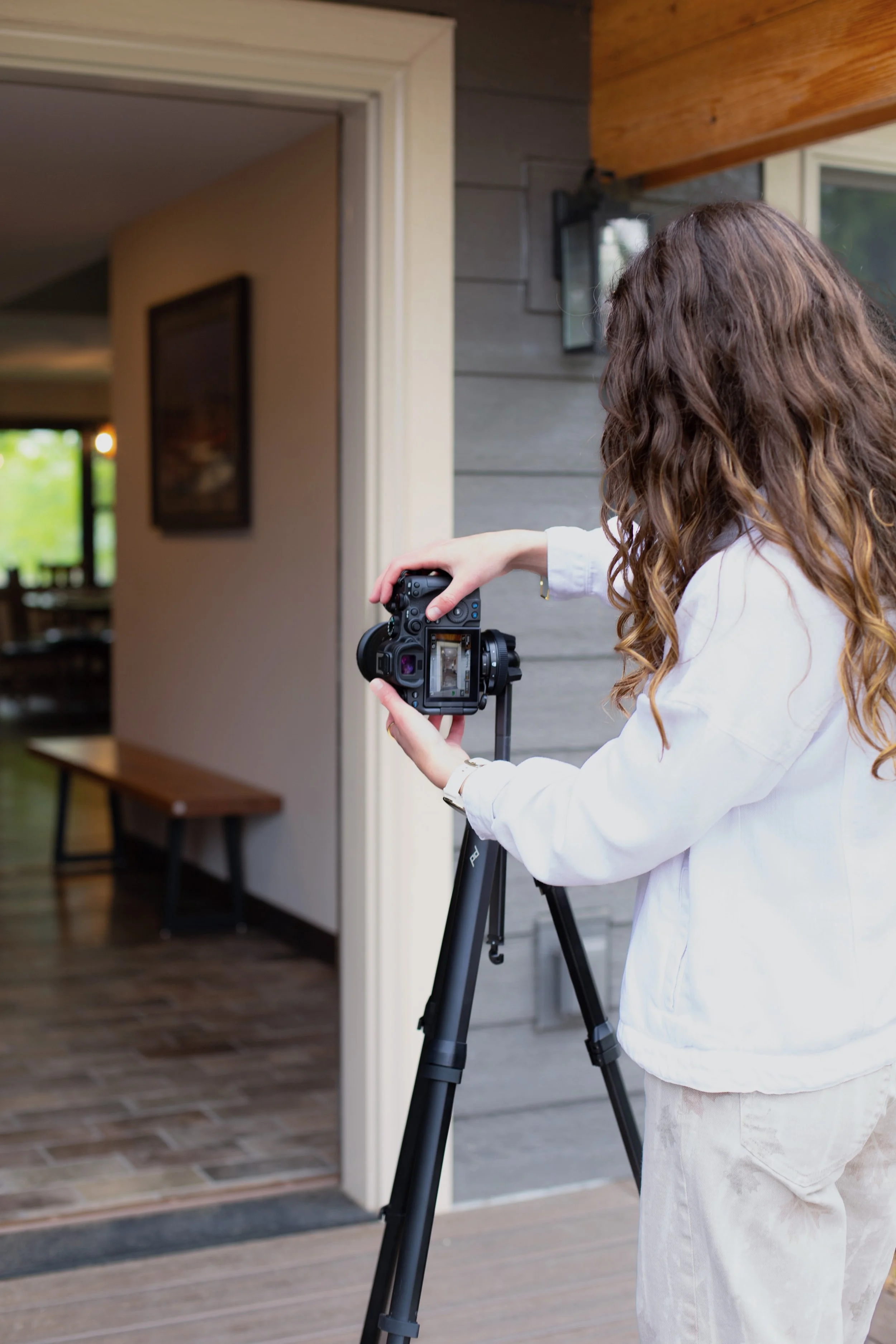 A woman with long, wavy hair using a Mirroless camera on a tripod outside a house, aiming the camera at the interior through an open door.