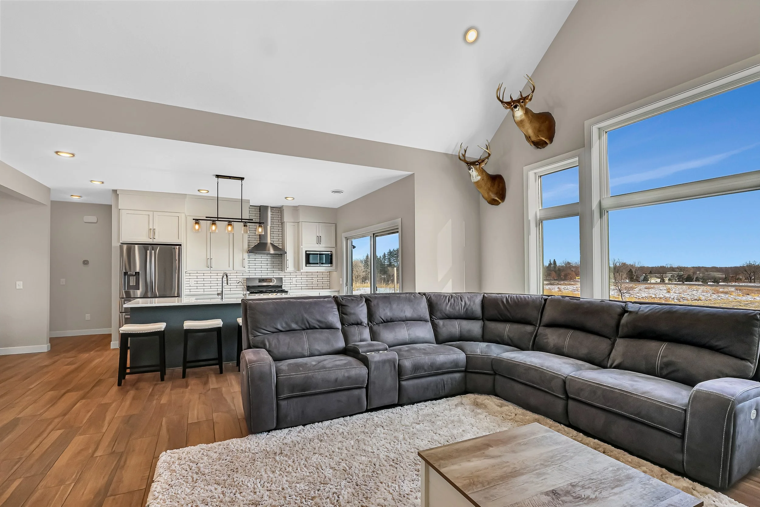 Living room with large sectional sofa, wood flooring, and a white shag rug, overlooking an open kitchen with white cabinets, stainless steel appliances, and a view of a snowy landscape through large windows, with mounted deer head decor.