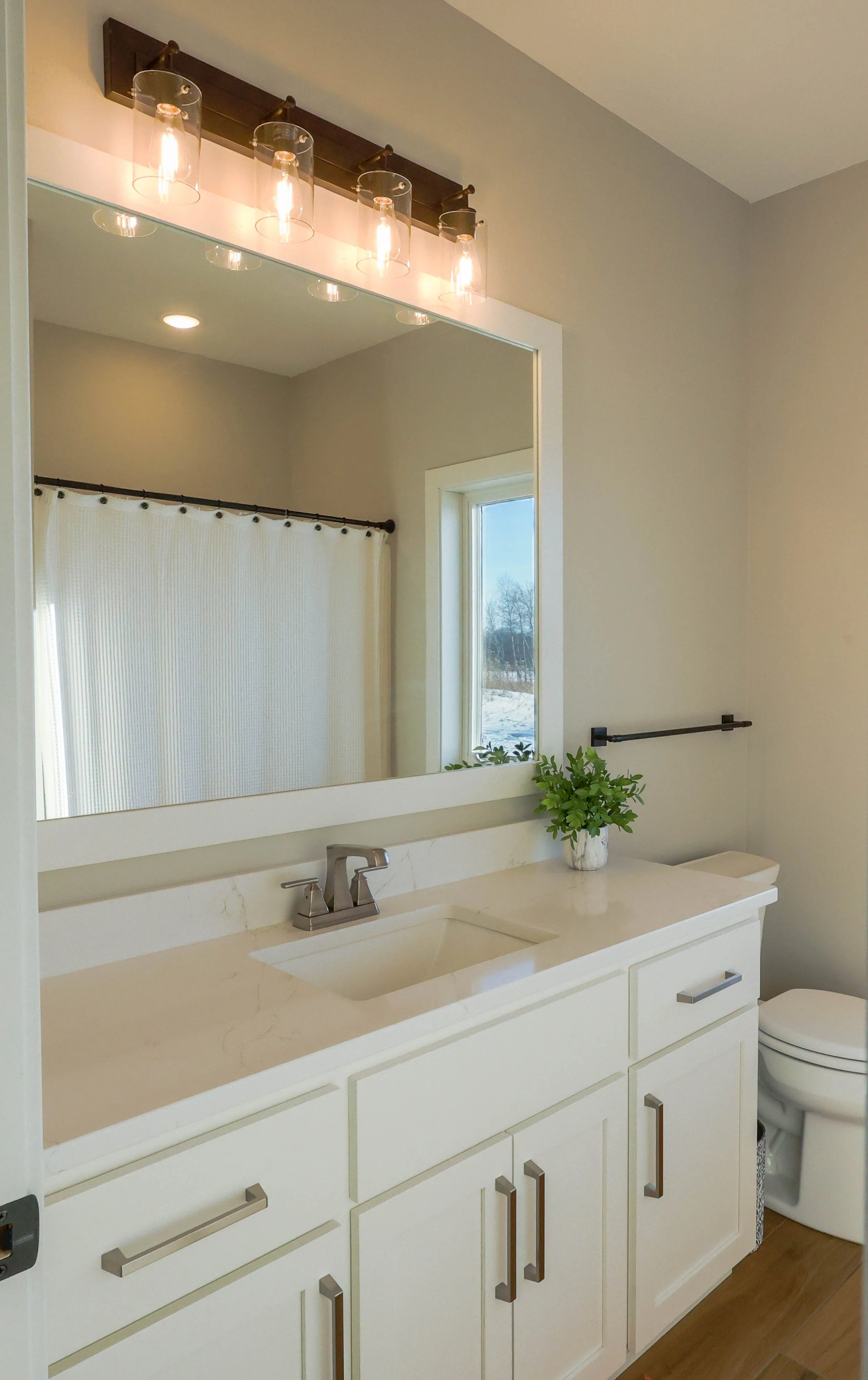 Bathroom with white vanity, sink, silver faucet, large mirror, window, light fixture with five glass shades, potted plant, and toilet.