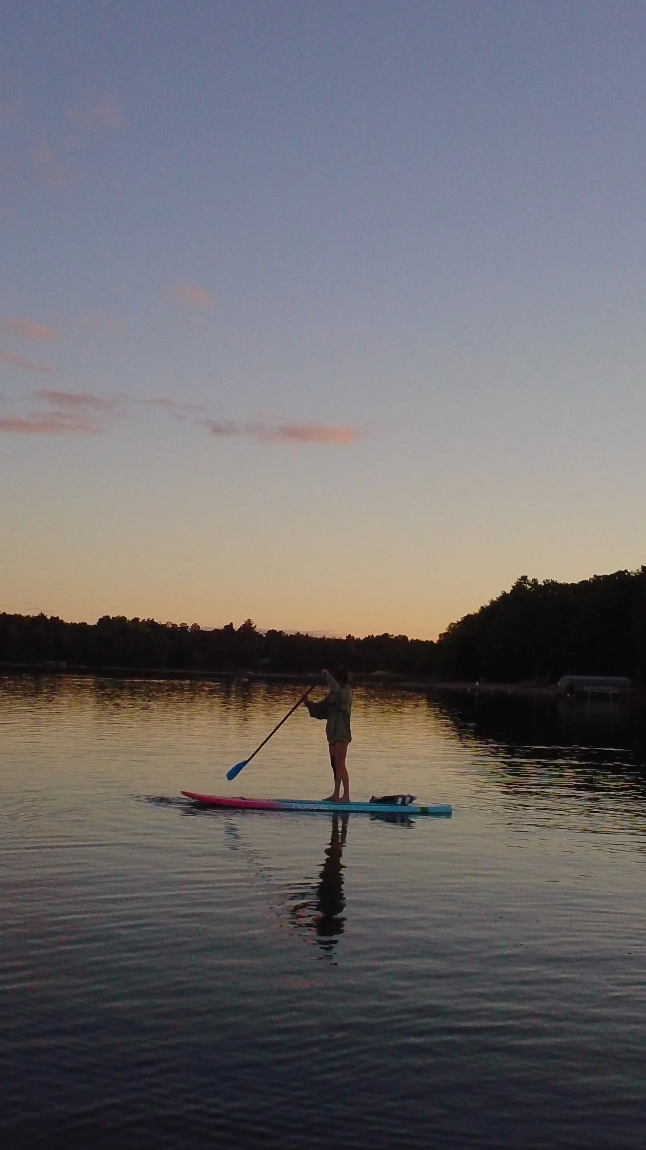 Person paddleboarding on calm water during sunset, with trees in the background and a sky with soft pastel clouds.