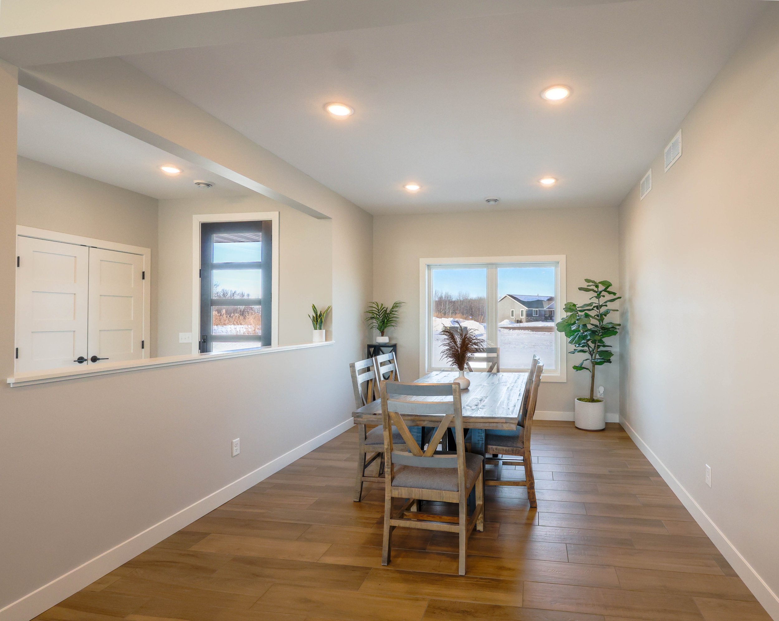Bright dining area with natural light, wooden table surrounded by chairs, potted plants, large windows showing a snowy neighborhood, recessed lighting, and white walls.