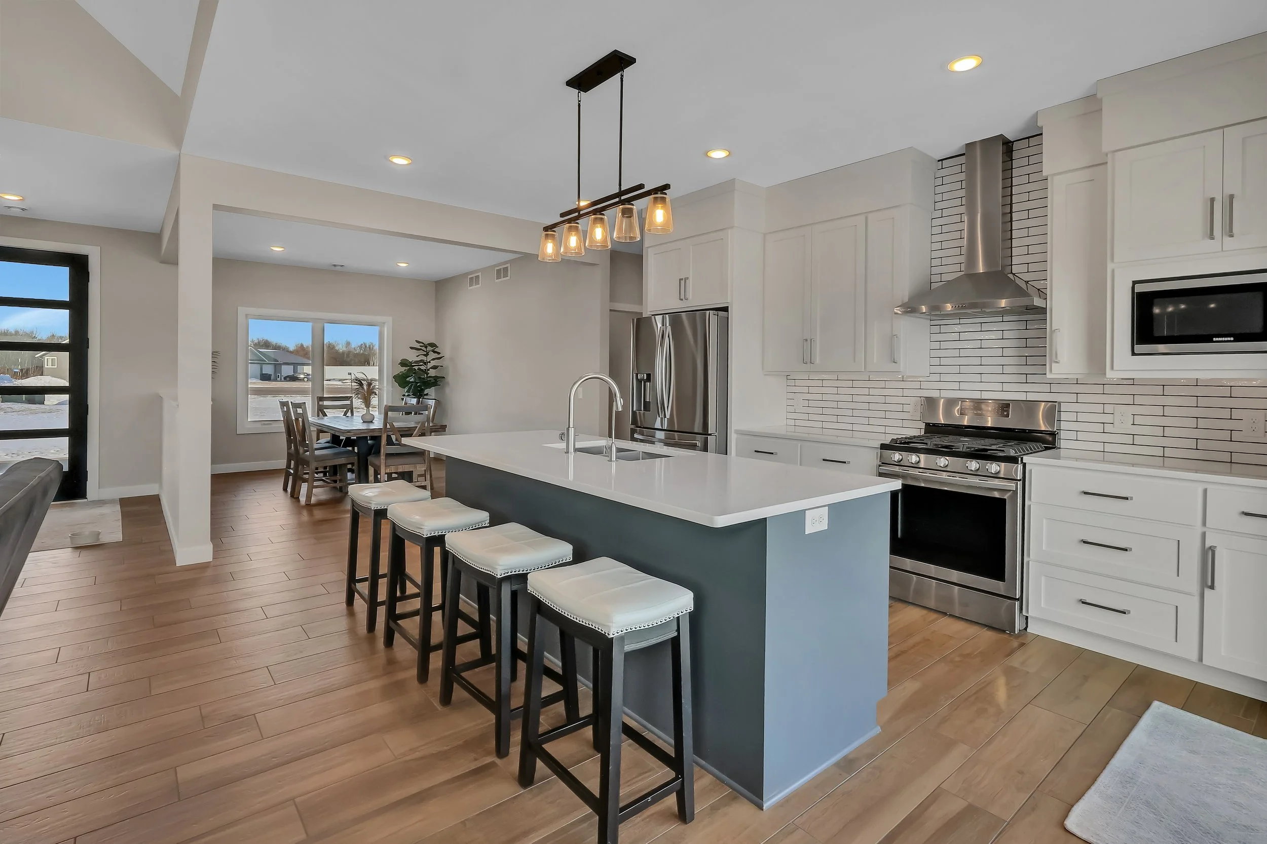 Modern kitchen with white cabinets, stainless steel appliances, a large island with a white countertop and four barstools, and a dining area with a wooden table and chairs near a window.