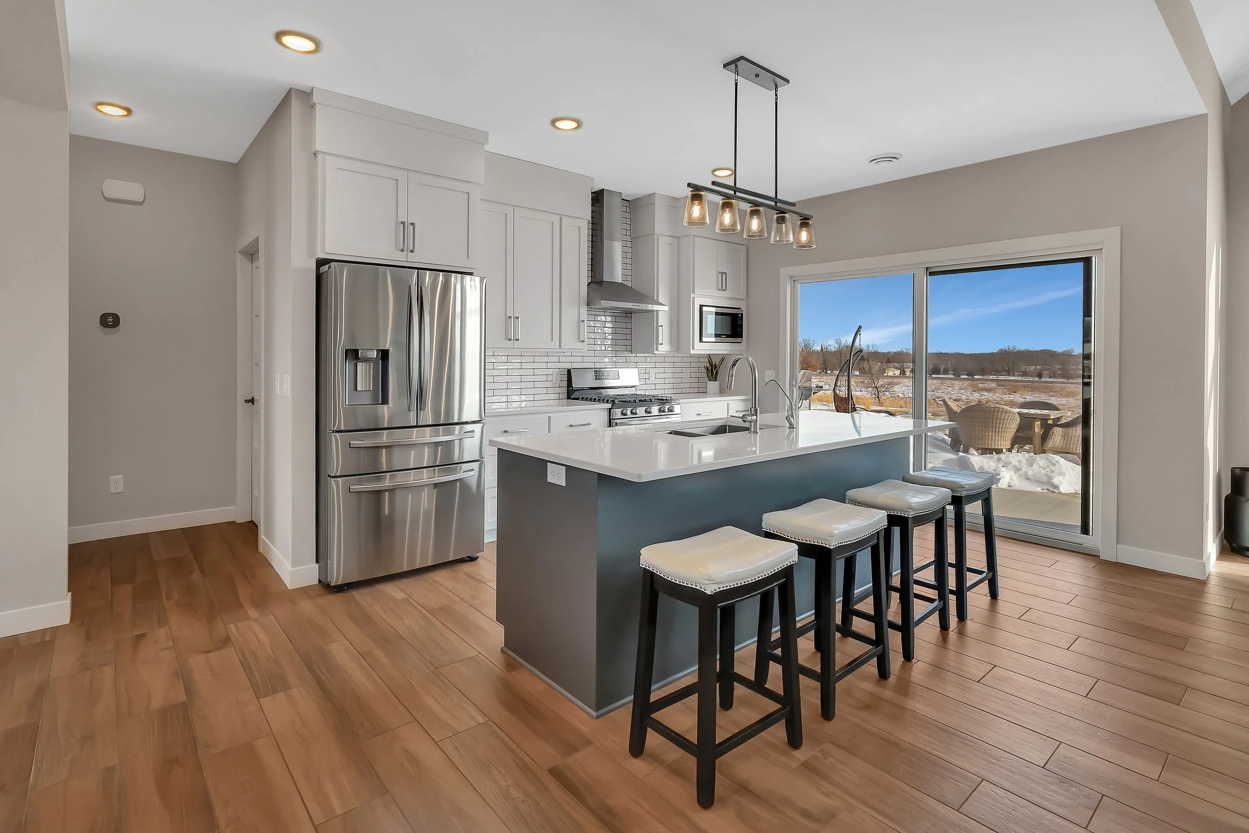 Modern kitchen with white cabinets, stainless steel refrigerator, stove, microwave, and a large window overlooking a snow-covered outdoor patio with outdoor furniture. An island with four bar stools and pendant lighting.