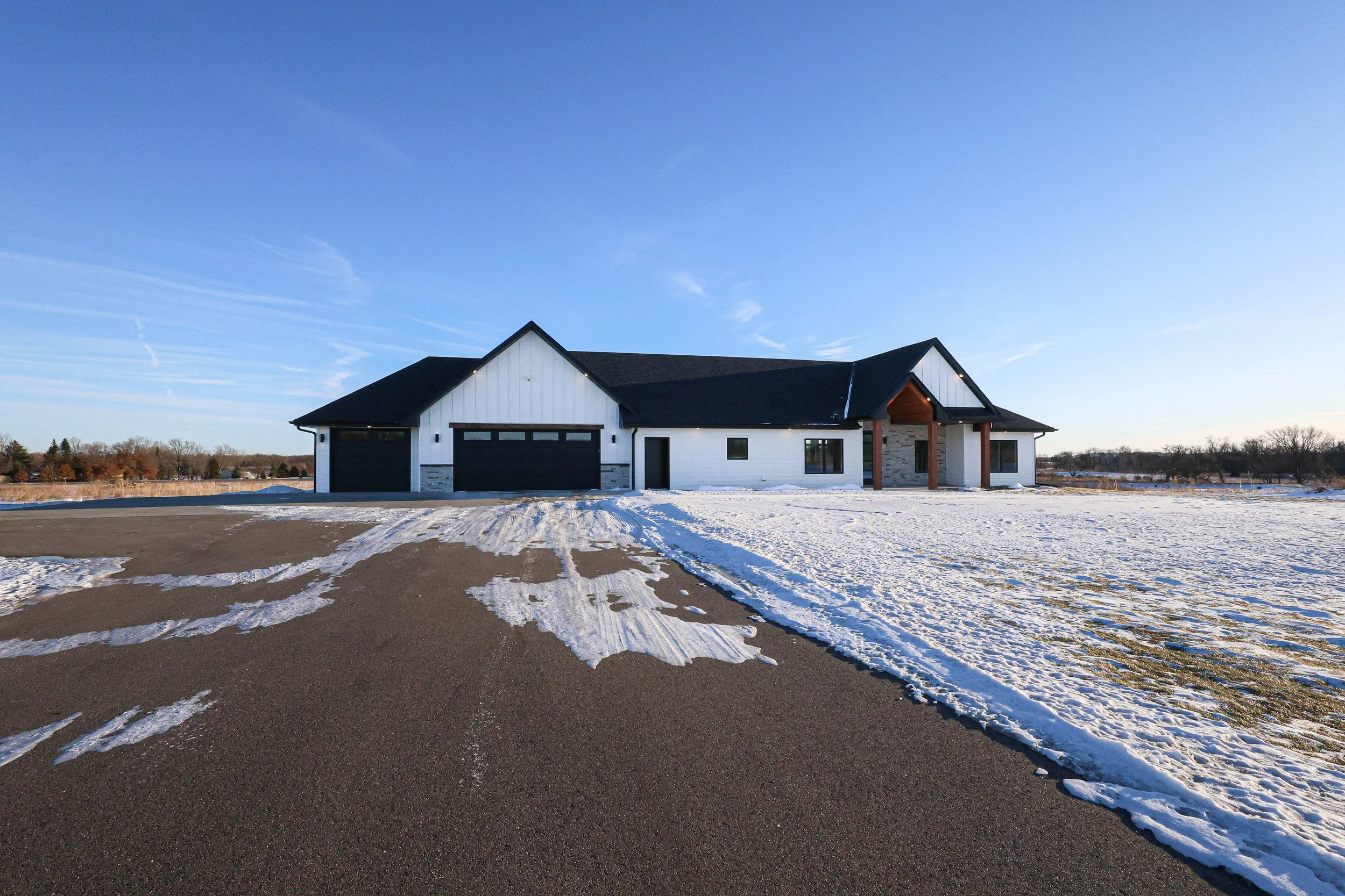 A modern house with a black roof and white walls, surrounded by snow, with a driveway in the foreground and clear blue sky above.
