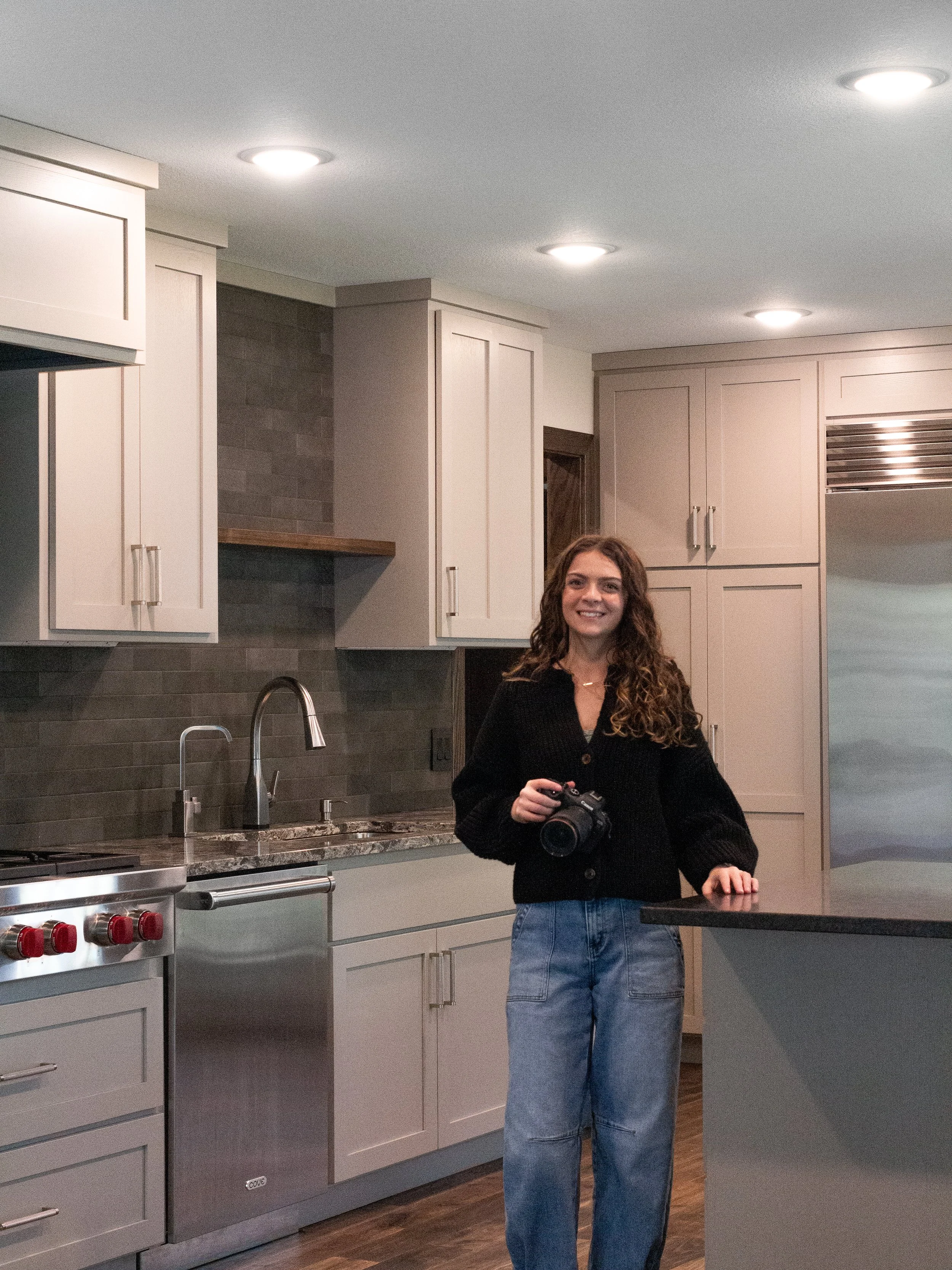A woman with long, curly hair holding a camera stands in a modern kitchen with white cabinets, a gray backsplash, and a stainless steel cove dishwasher and a wolf gas cooktop. She is smiling and wearing a black sweater and blue jeans.