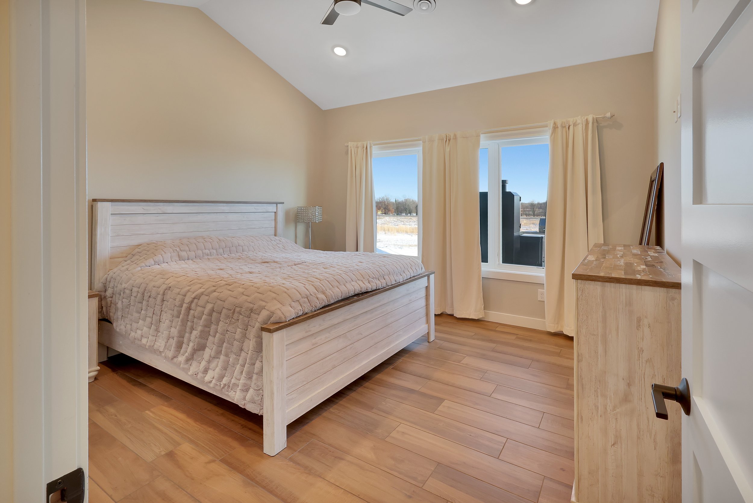 A bedroom with a wooden bed, beige quilt, and cream curtains on large windows showing a wintery outdoor landscape.