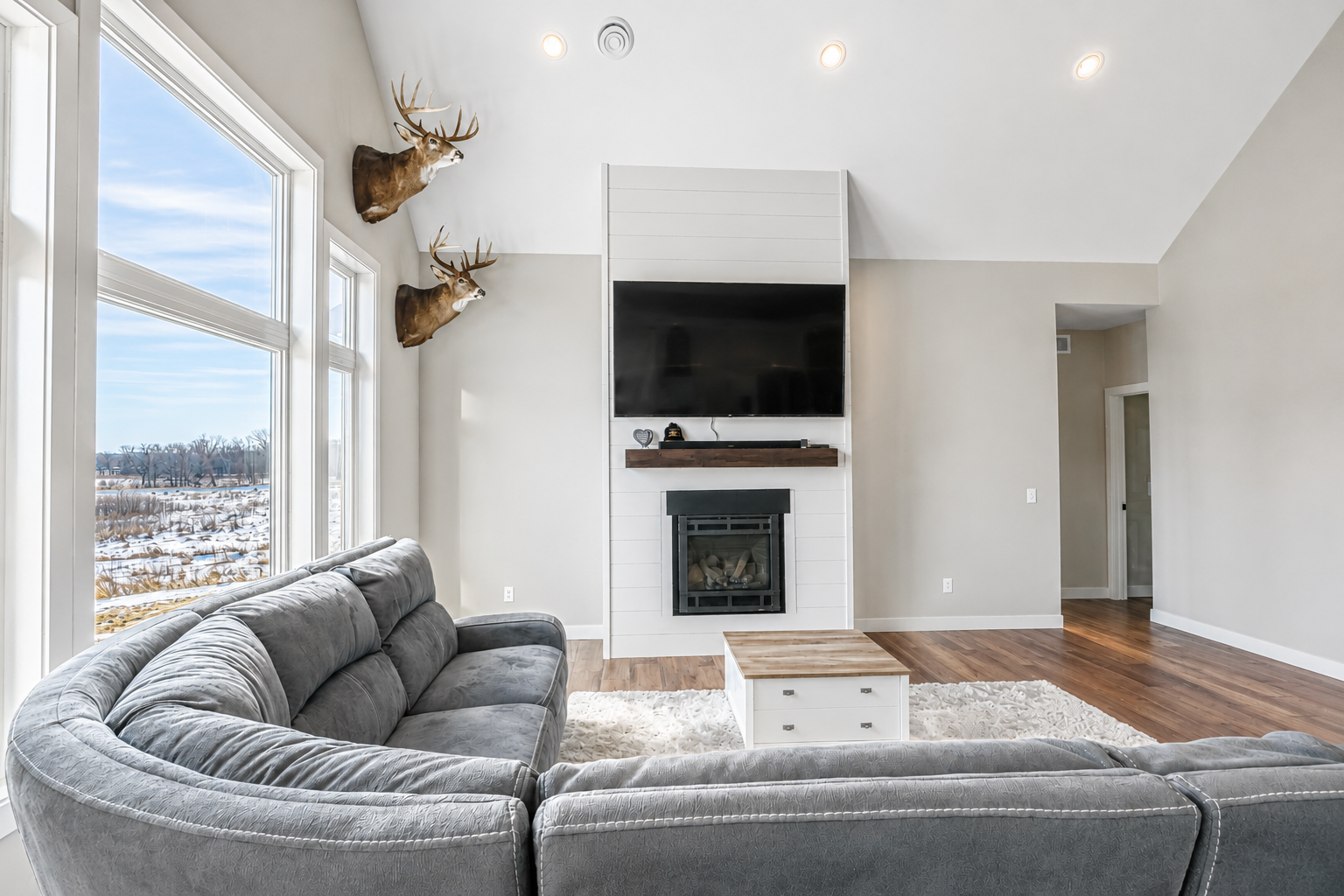 Living room with large windows, a grey sectional sofa, a white coffee table, a modern fireplace, a mounted TV, and deer head wall mounts on the wall near the windows.