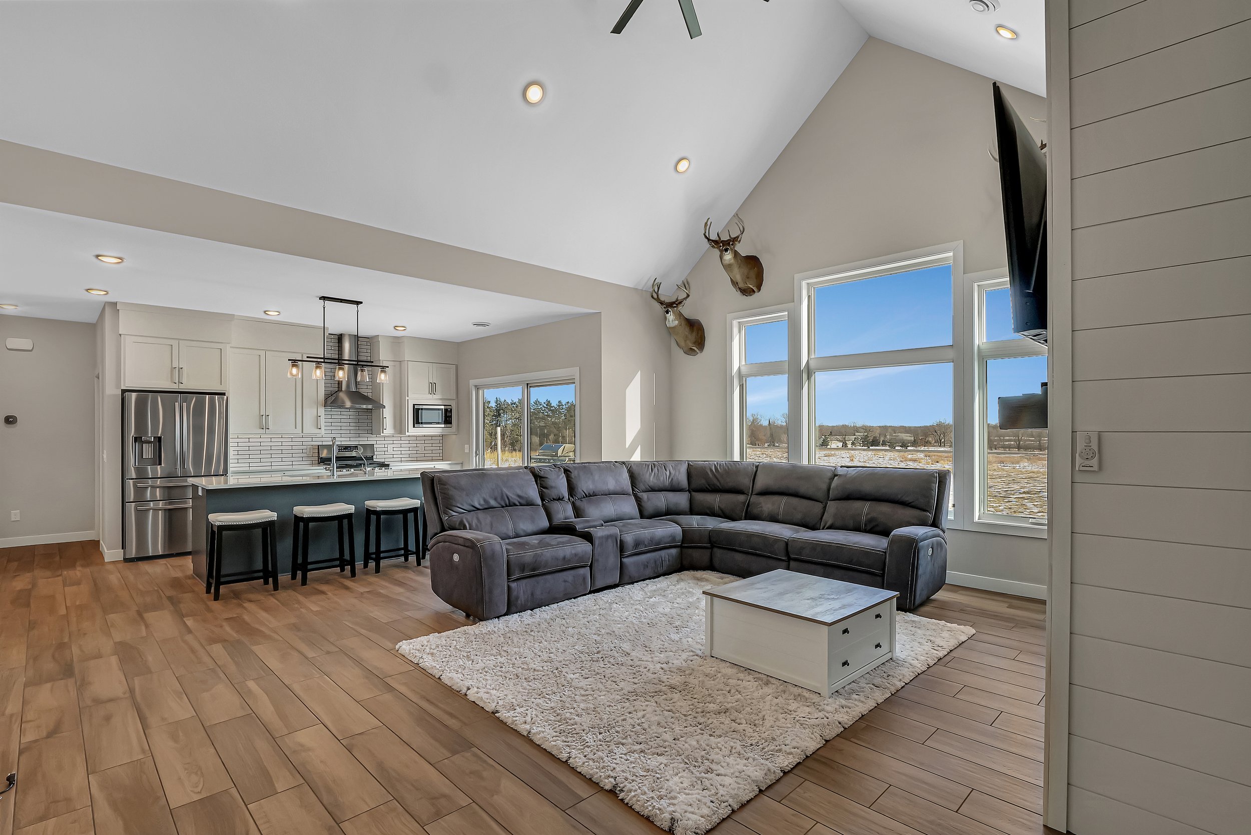 Living room with large window, gray sectional sofa on a cream rug, white coffee table, and view of the outdoors. Kitchen in background with white cabinets, stainless steel appliances, and black bar stools.