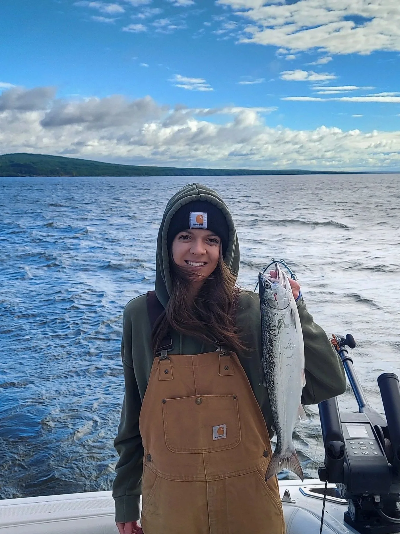 A woman in a hoodie and Carhartt overalls holding a lake trout on a boat on lake superior, with a hilly landscape and blue sky with clouds in the background.