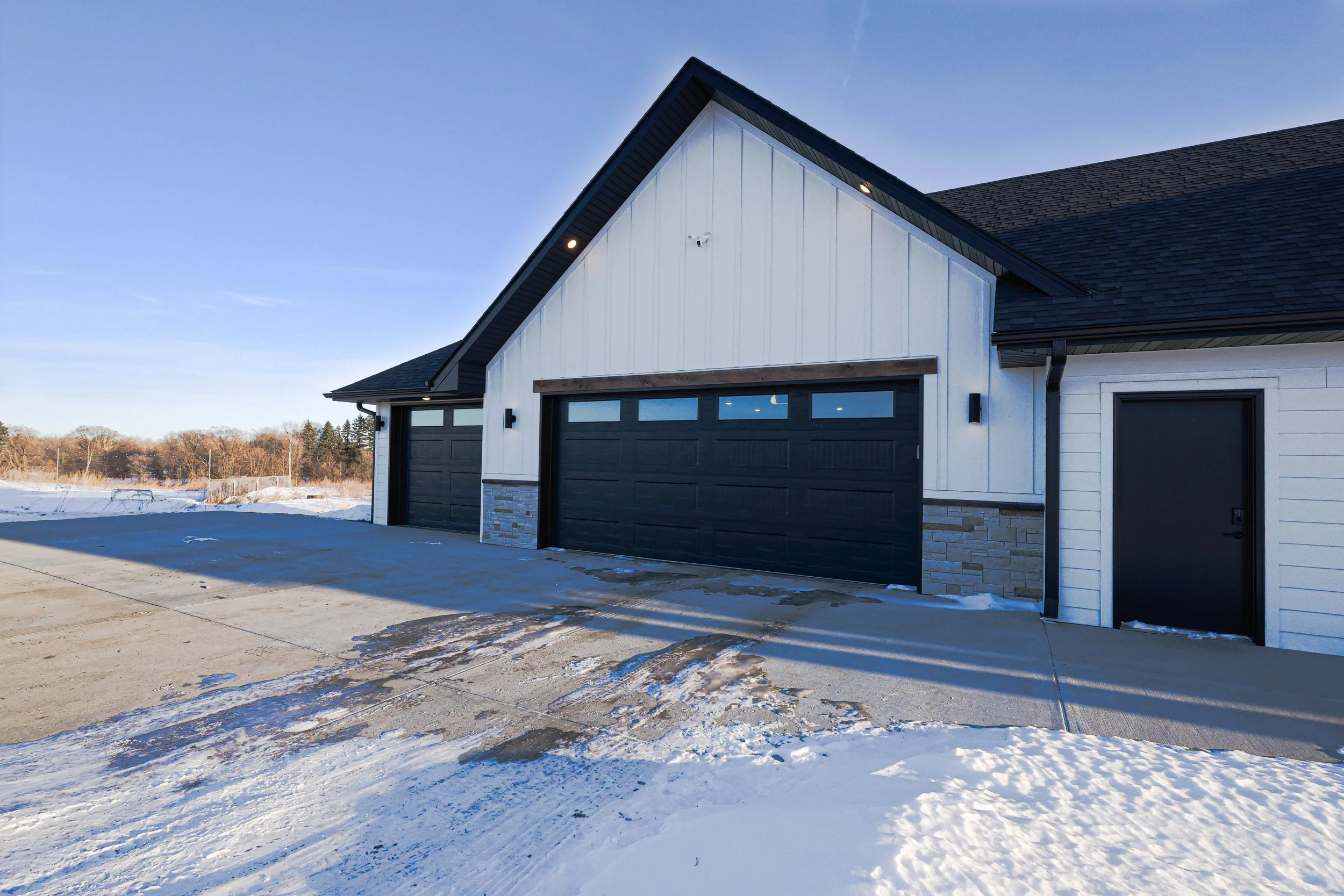 A modern house with a dark garage door, white siding, and a sloped roof, photographed on a snowy day with clear blue sky.