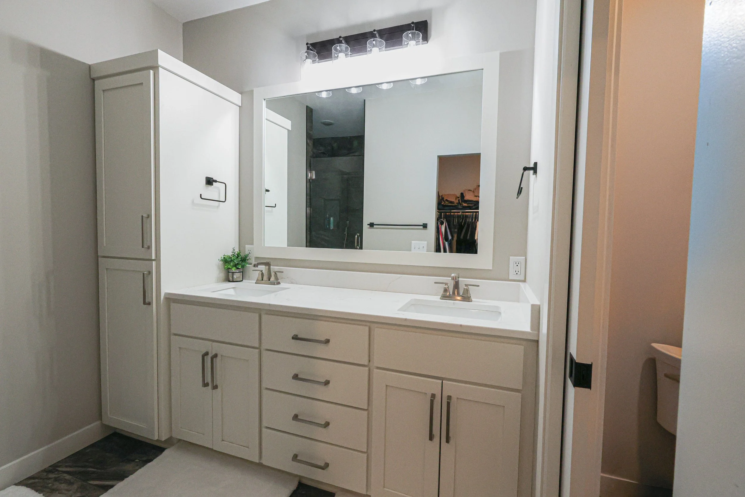 Bathroom vanity with a large mirror, white cabinets, and a double sink, with a small potted plant and soap dispenser on the countertop.