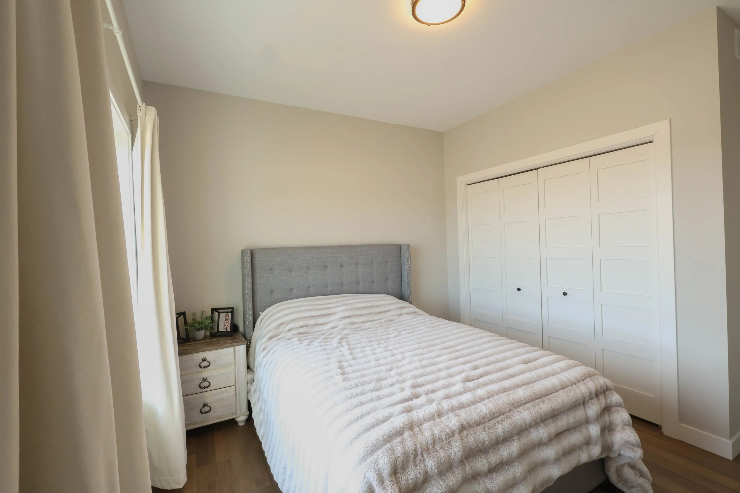 A bedroom with a bed covered in a beige and white striped quilt, a gray upholstered headboard, a white nightstand with a small plant, wireless speaker, and framed photo, cream-colored curtains on a window, and white closet doors with six panels, in a