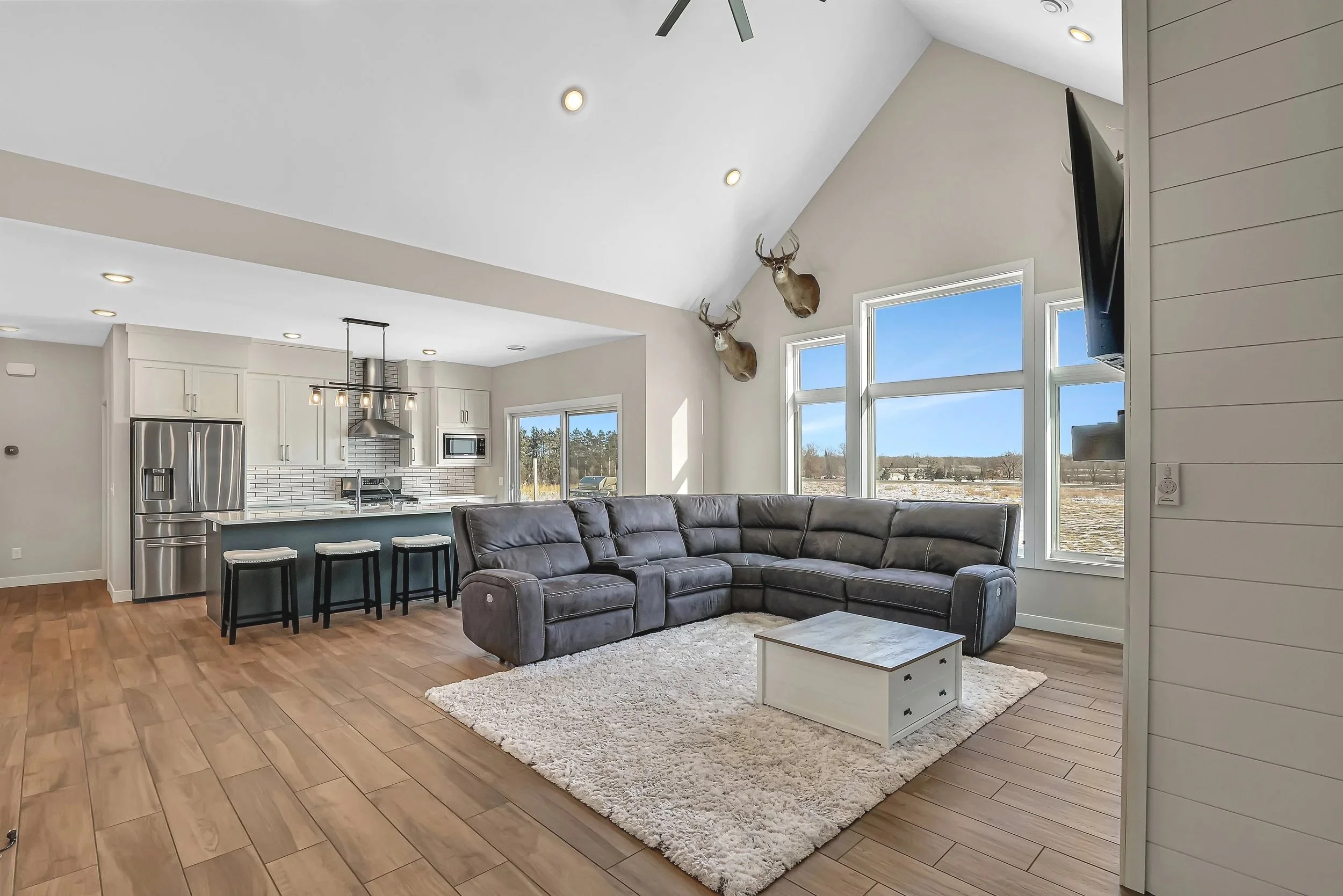 Living room with gray sectional sofa on a white rug, large windows showing a blue sky and trees, mounted deer heads on the wall, modern kitchen in the background with white cabinets and stainless steel appliances, hardwood floors, ceiling fan, recessed lighting, and a hanging light fixture over the kitchen island.