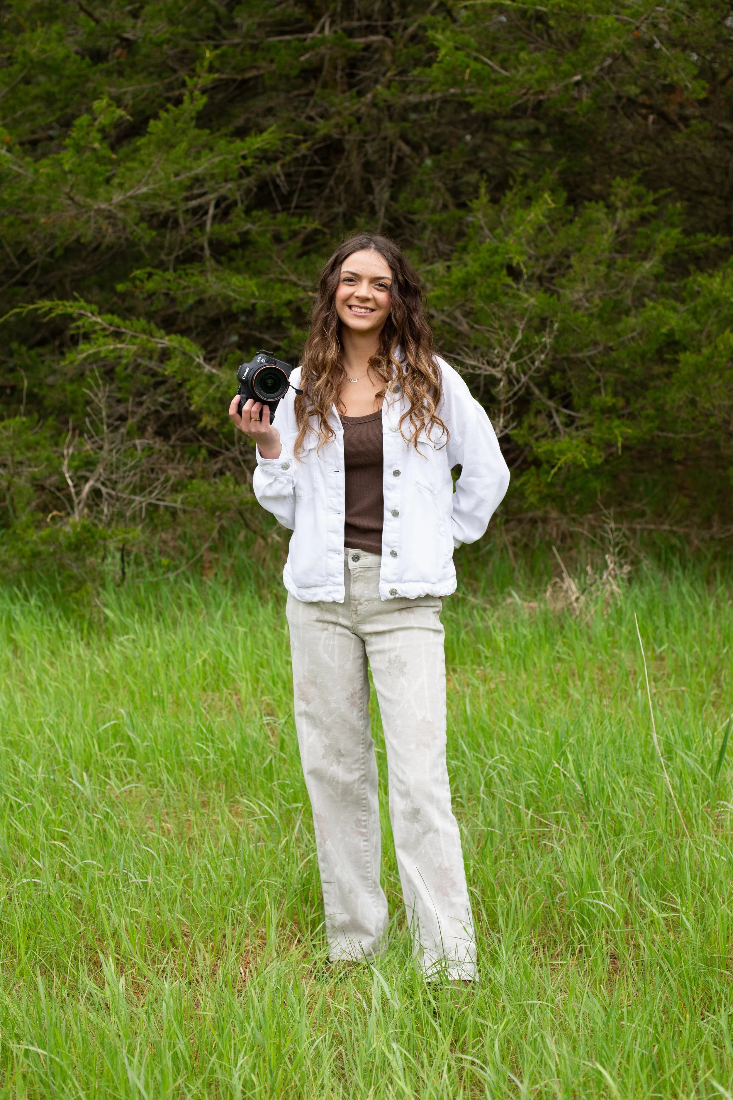 A woman standing in tall green grass outdoors, holding a camera, smiling, with trees in the background.