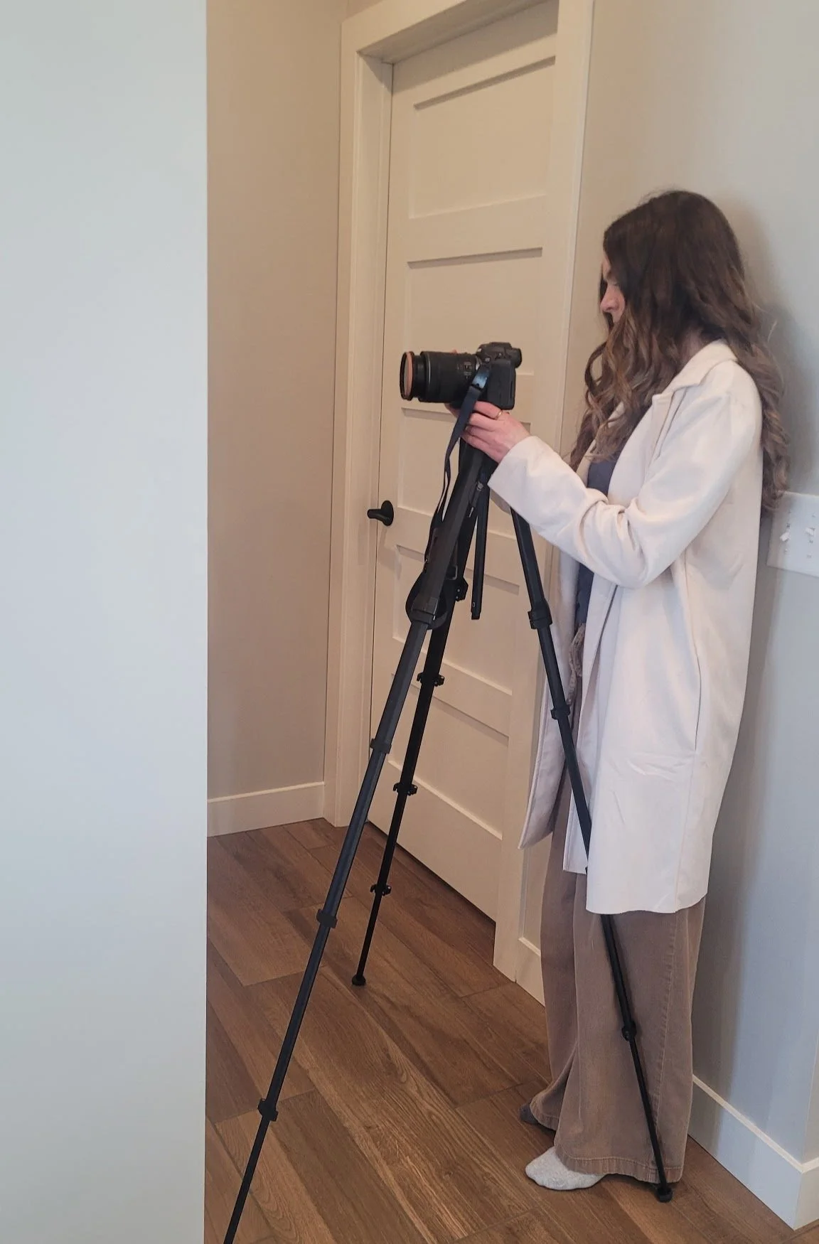 A woman with wavy brown hair wearing a beige coat and brown pants standing on a wooden floor, looking at a camera mounted on a tripod next to her inside a room with white walls and a white door.