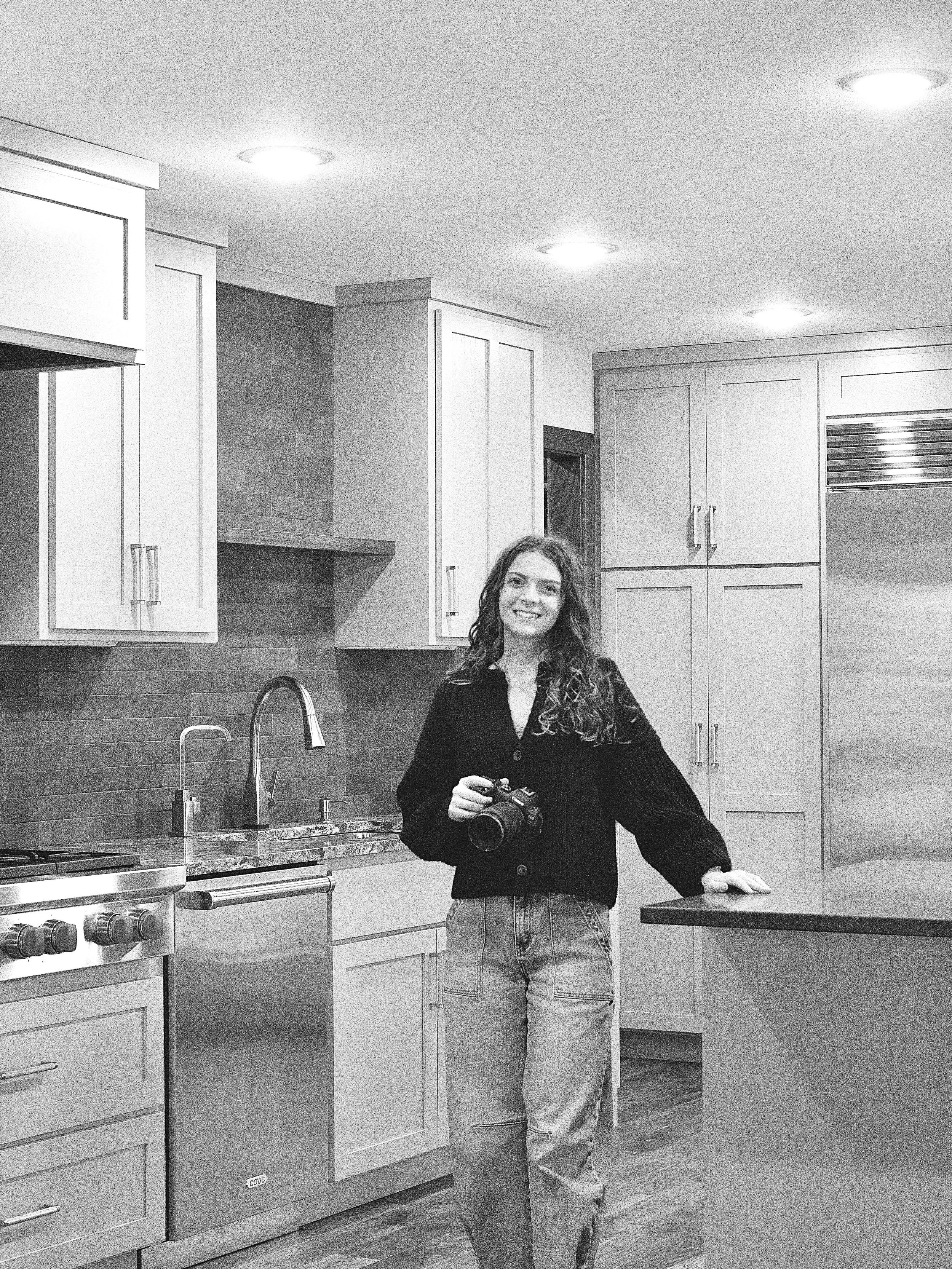 A woman with long curly hair, smiling, standing in a modern kitchen holding a camera in her right hand. She is wearing a dark button-up shirt and light jeans. The kitchen has white cabinets, a stainless steel stove, a sink, and overhead lighting.