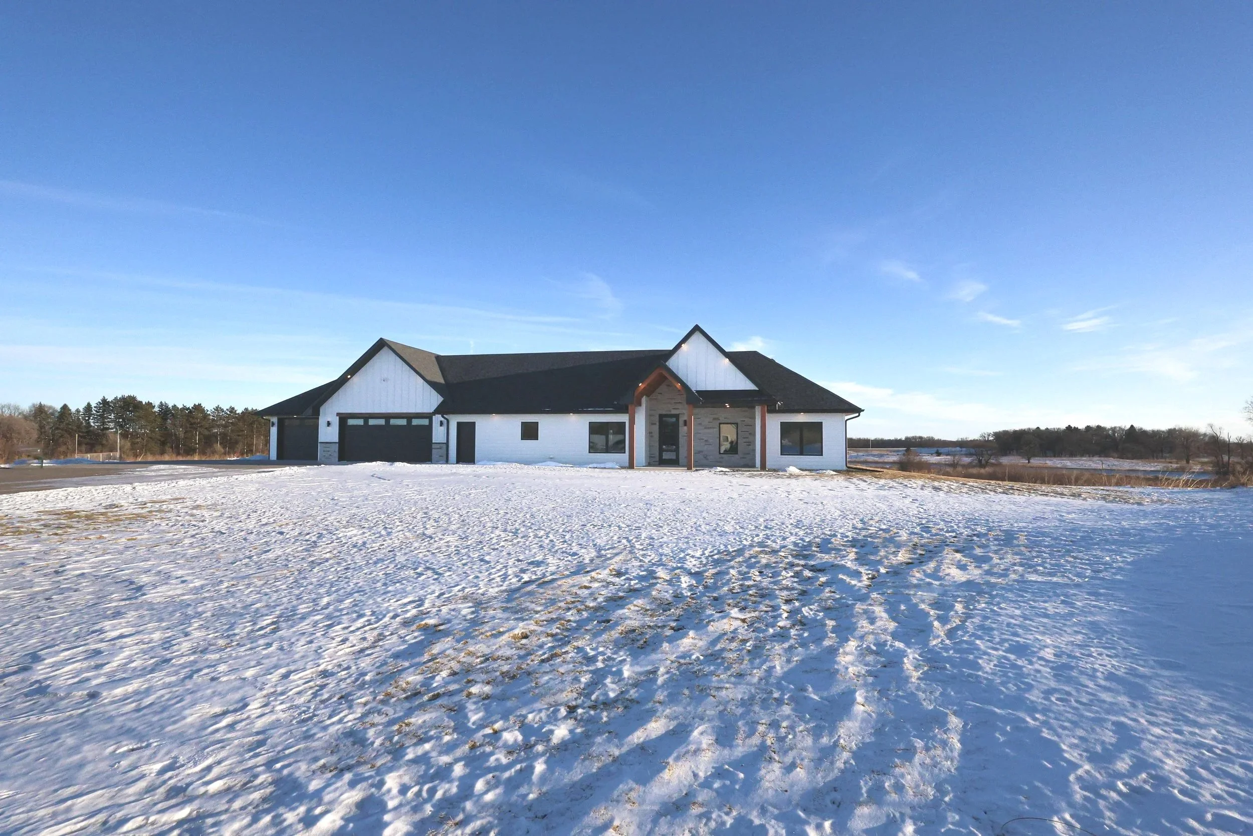 A house with a modern design, white exterior walls, large windows, and a dark roof in a snowy landscape under a clear blue sky.