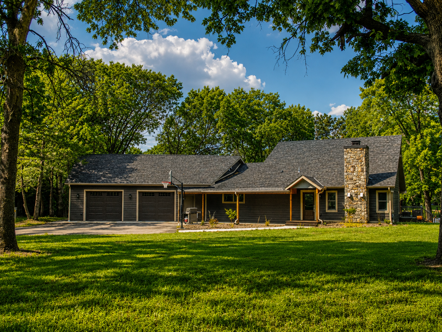 A modern house with black siding, a stone chimney, a two-car garage, a front porch, and a basketball hoop in the driveway, surrounded by green trees and grass on a sunny day.