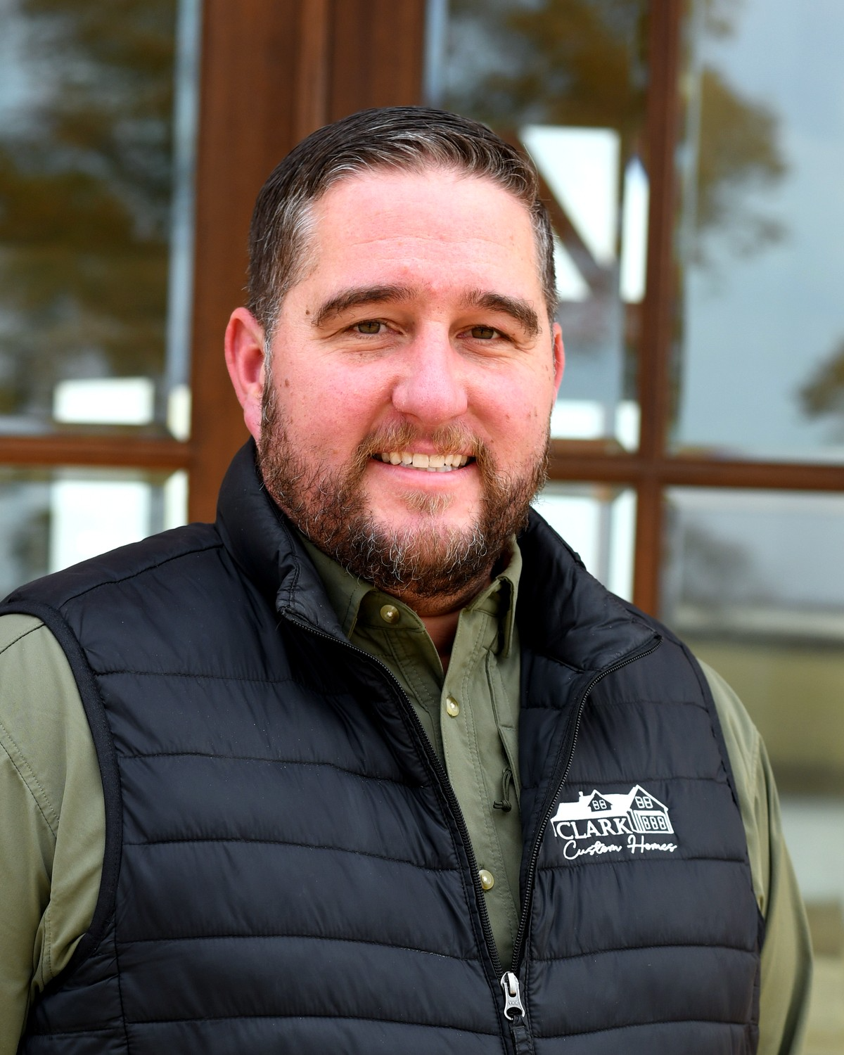 A man with short dark hair and a beard, smiling, wearing a black puffer vest over a green collared shirt, standing outdoors in front of a reflective glass building.