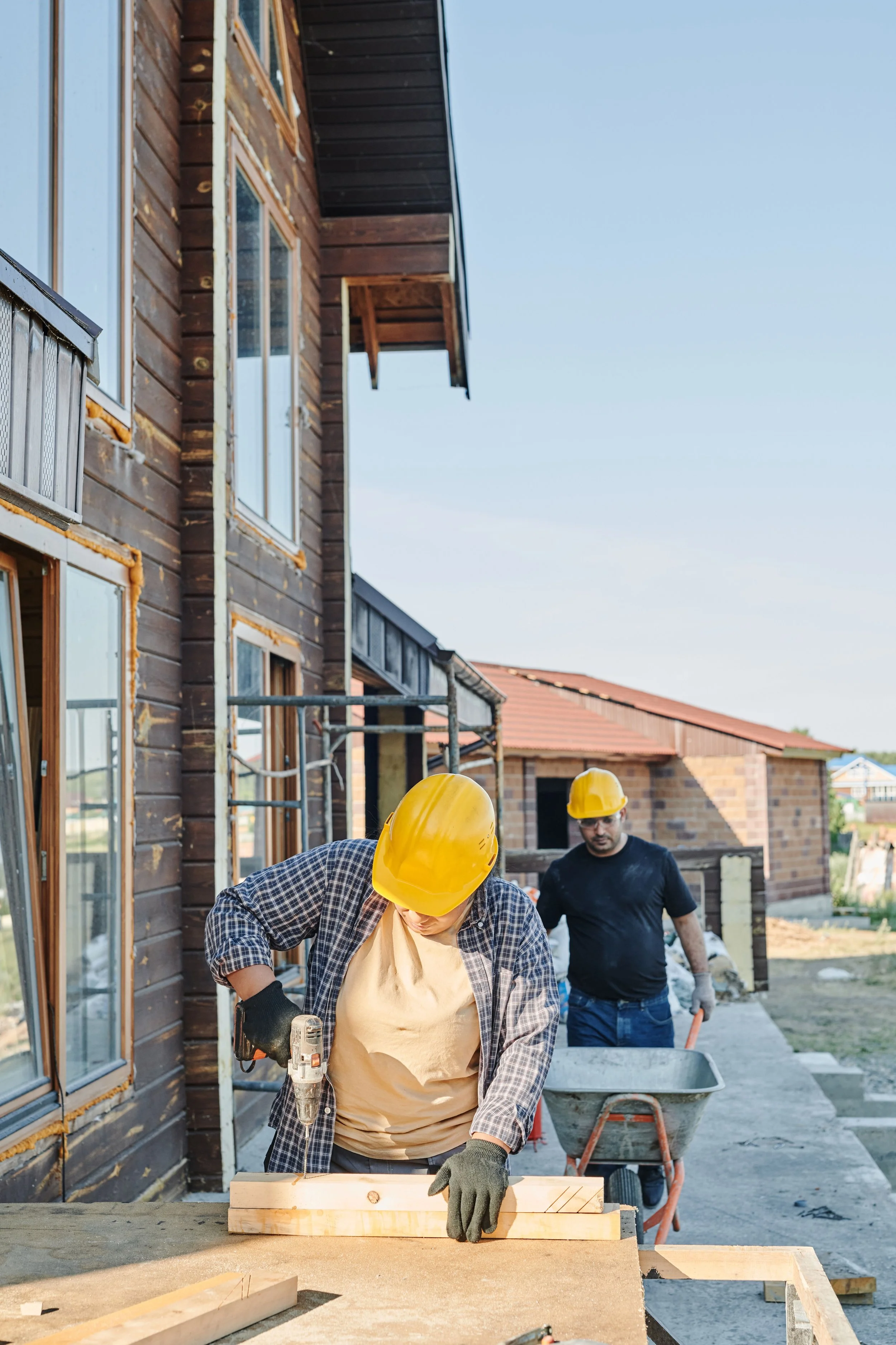 Construction workers in helmets working on building a wooden structure outside a house.