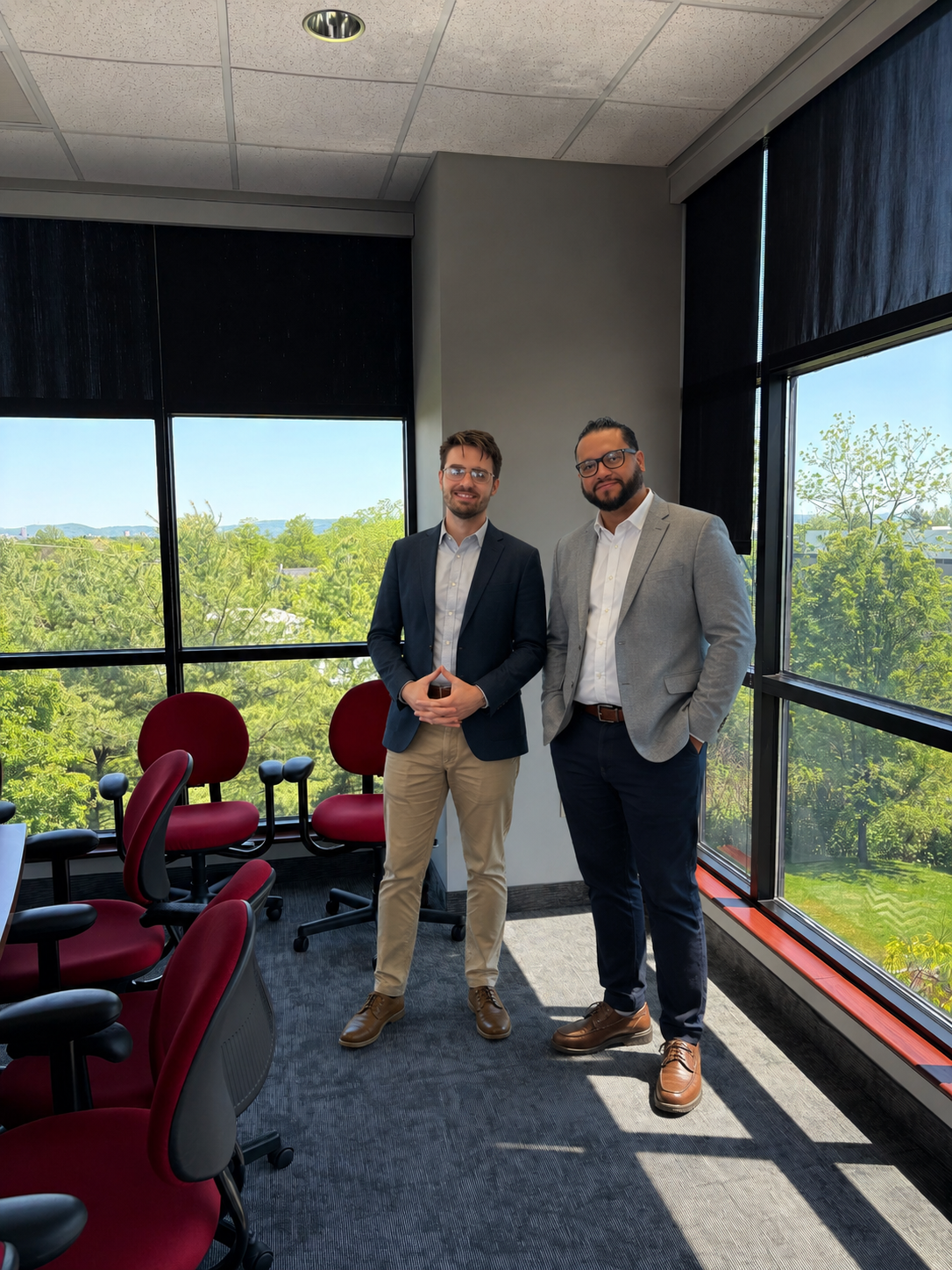 Two men in business attire standing in a conference room with large windows showing trees and a blue sky.