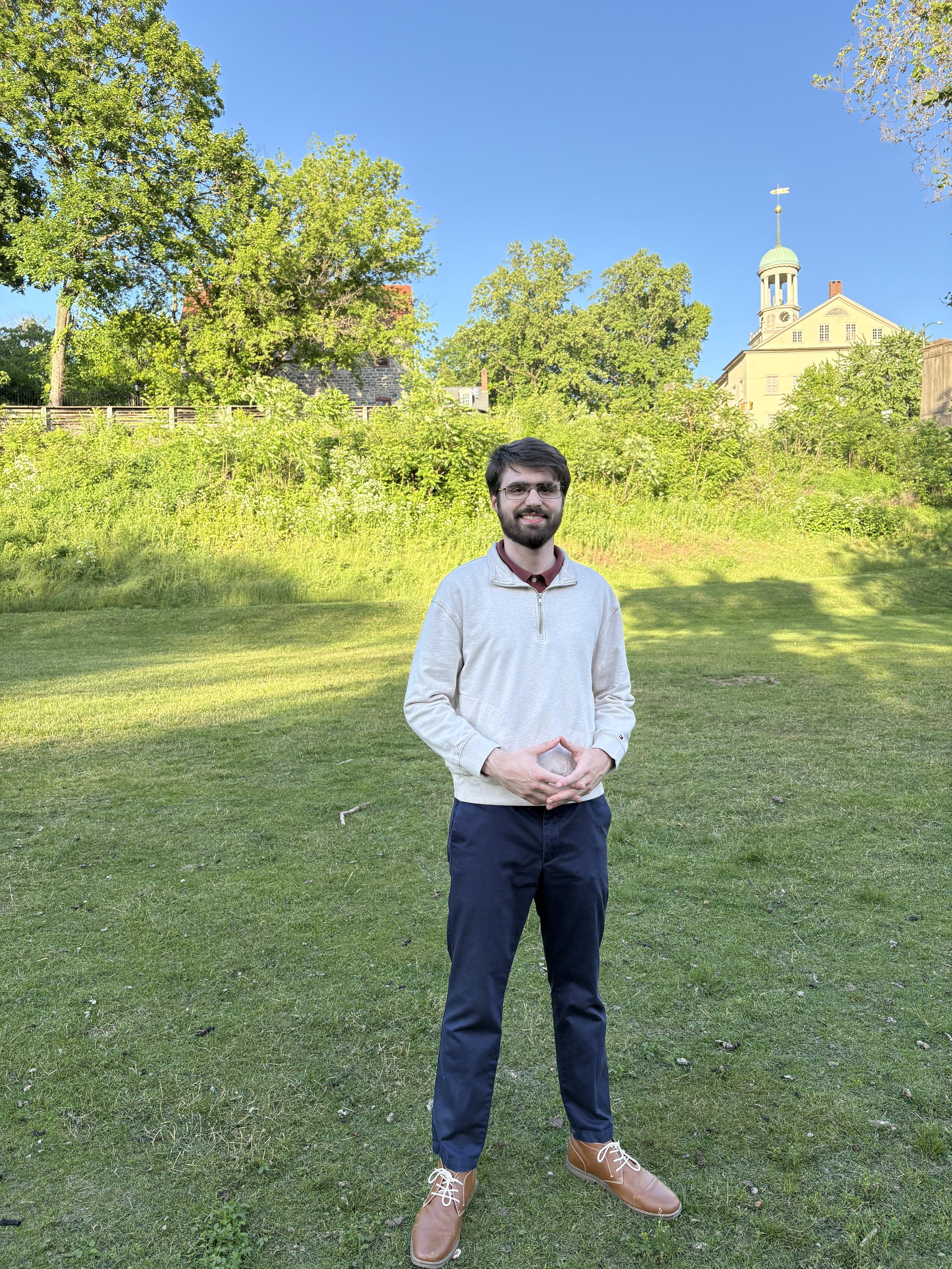 A man with a beard and glasses, wearing a light gray quarter-zip sweater, dark pants, and tan boots, standing on a grassy area with trees and a historic building with a clock tower in the background, on a clear sunny day.