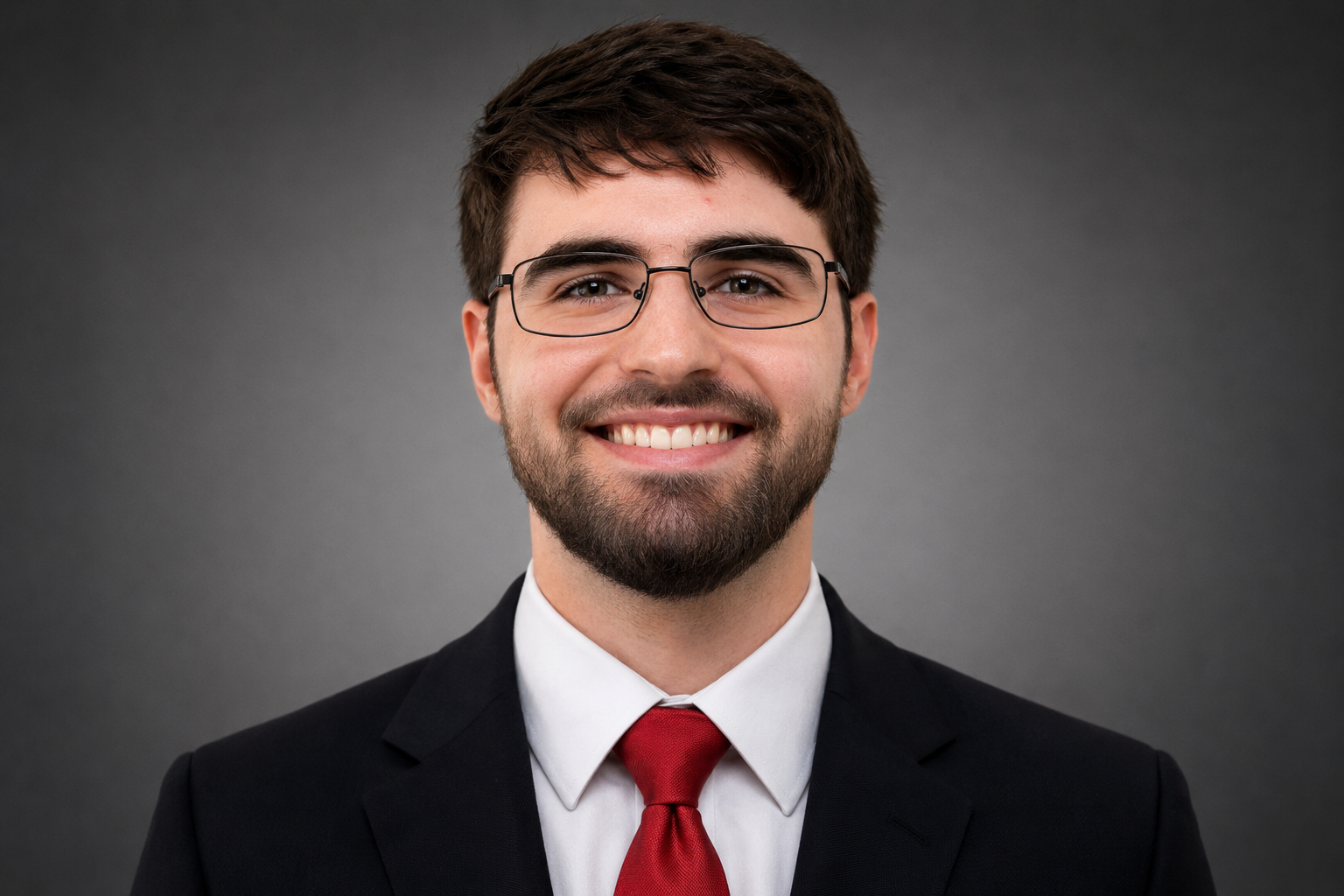 Professional headshot of a man with glasses, smiling, wearing a black suit, white shirt, and red tie against a gray background.