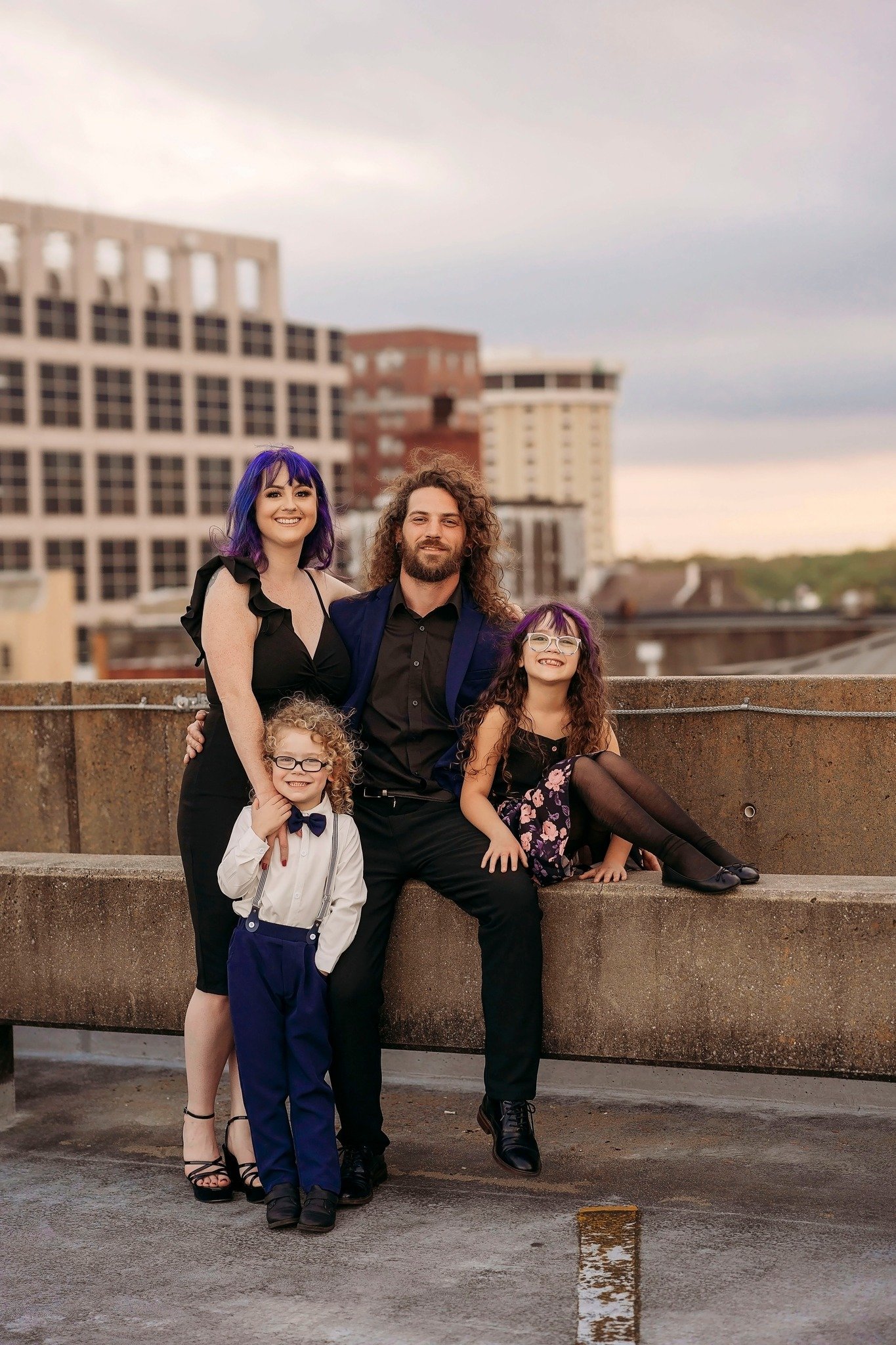 A family of five people including three children and two adults posing on an outdoor rooftop with city buildings in the background during sunset.