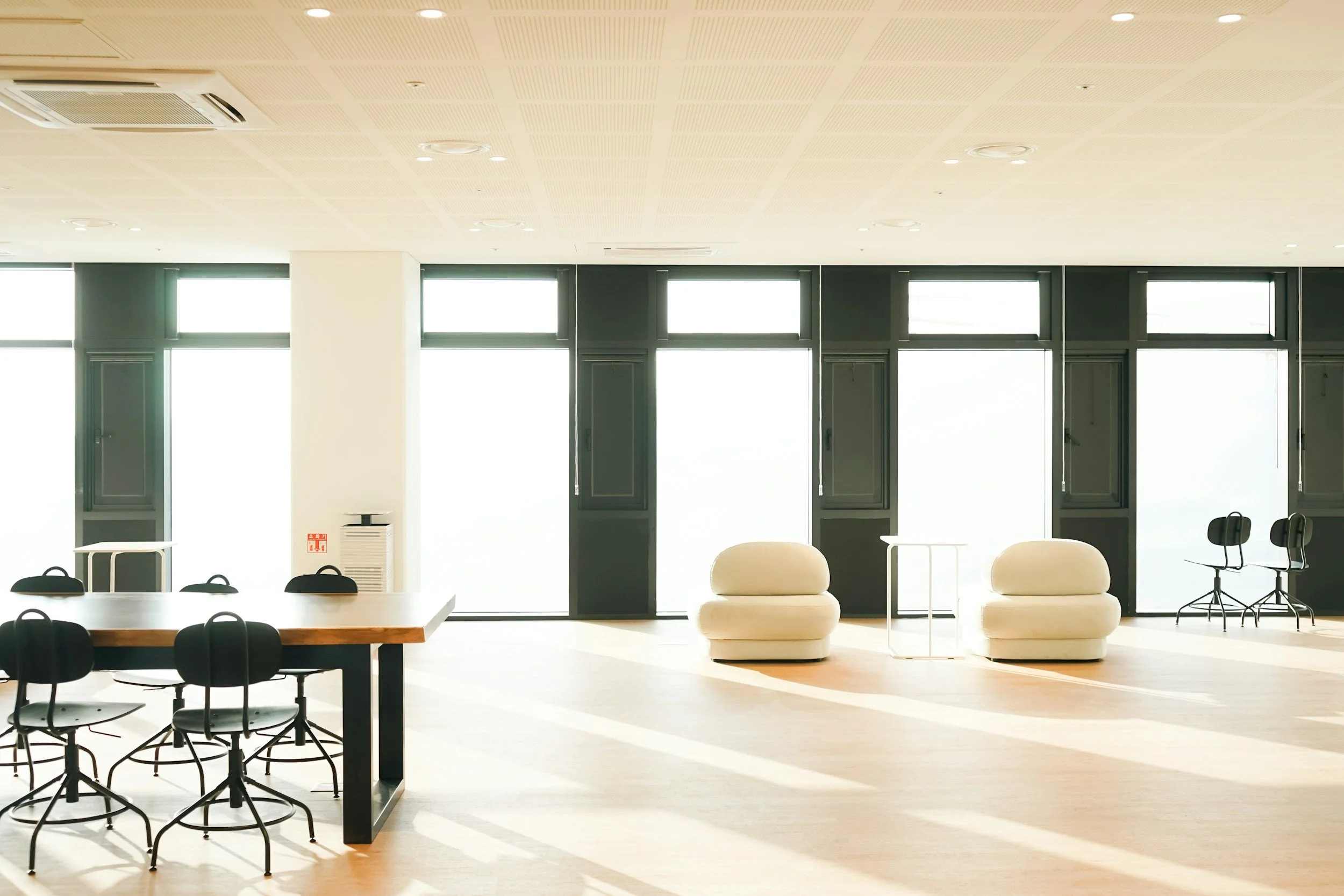 A modern bright office or conference room with large windows, white walls, and a high ceiling. There are tables and chairs, including a round table with black chairs, some beige sofas, and black office chairs, with sunlight streaming in and casting shadows on the light-colored floor.