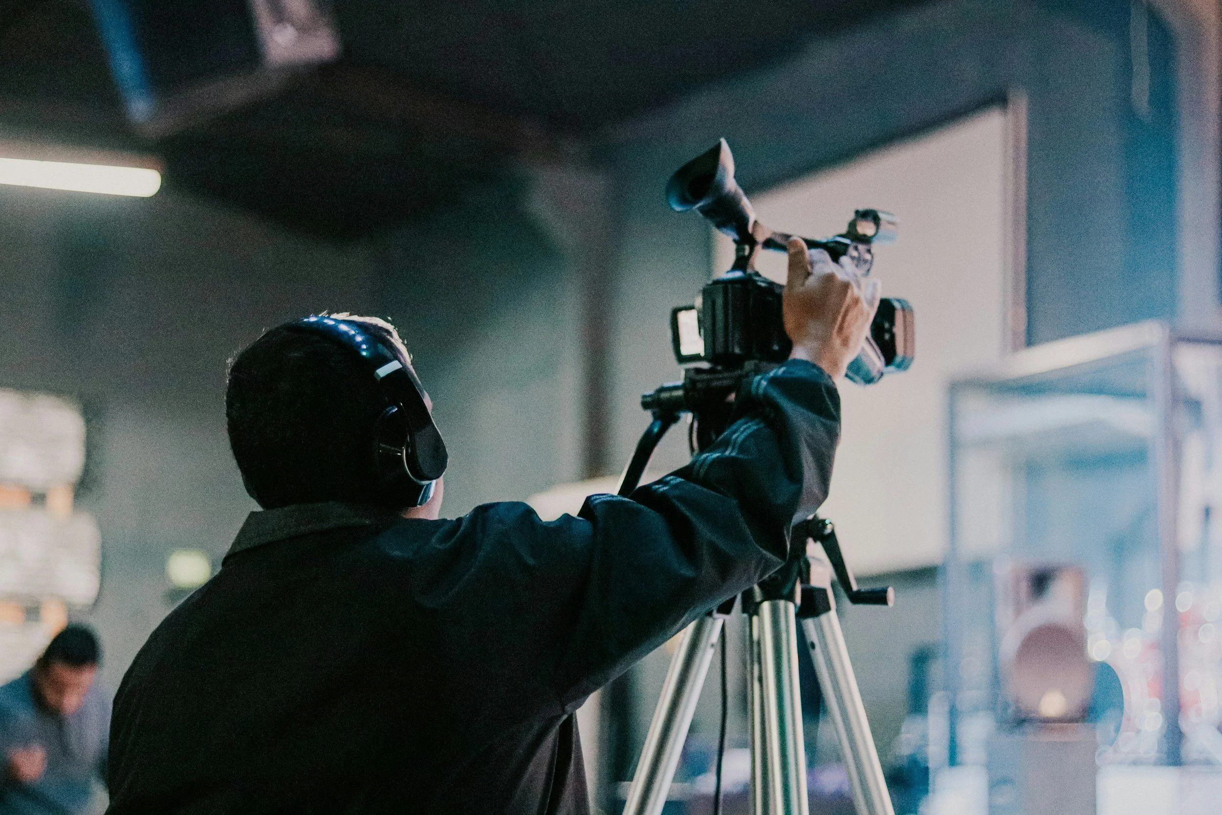 A person wearing a black jacket and helmet is adjusting a camera on a tripod indoors. The person is seen from behind.