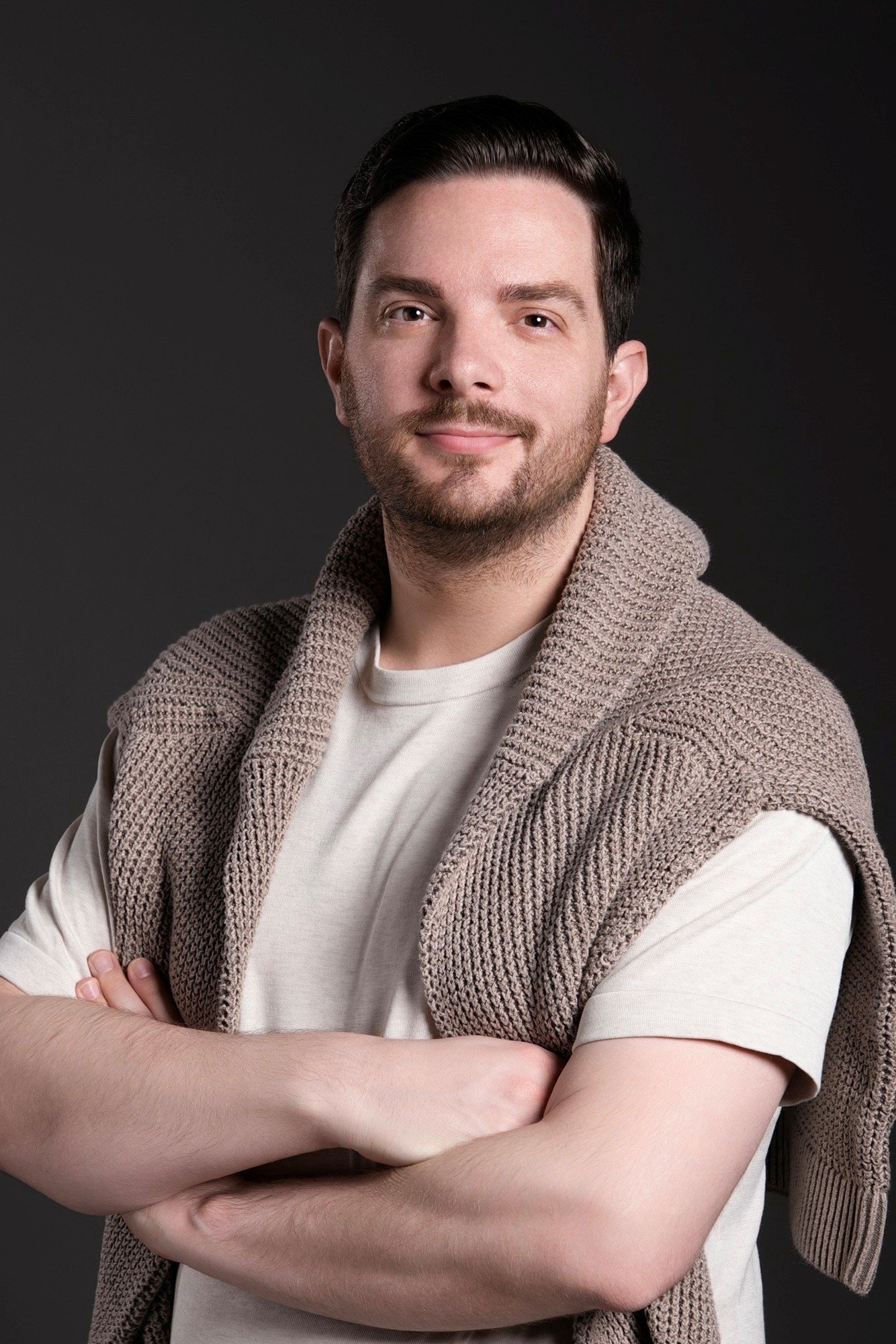 A man with dark hair, a beard, and light skin, standing with his arms crossed, wearing a beige knit sweater draped over a cream T-shirt, against a dark background.