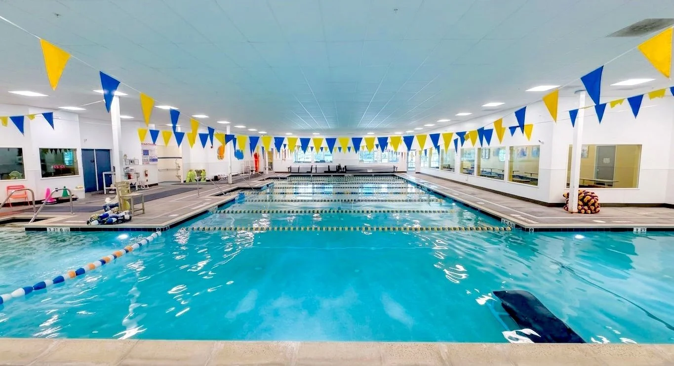 Indoor swimming pool with lane dividers, yellow and blue pennant banners, and windows along the wall.